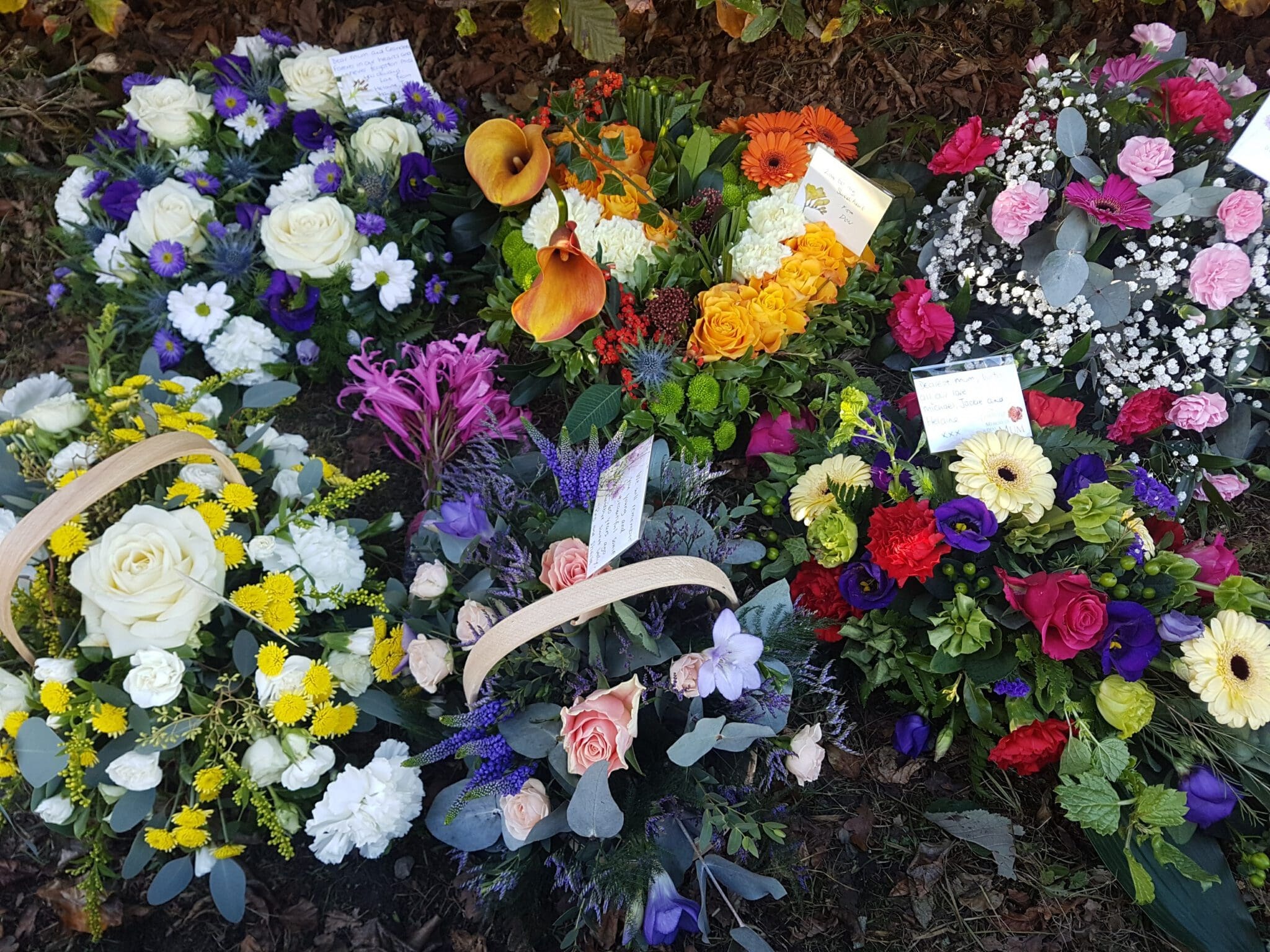 A variety of colorful floral arrangements, including roses, lilies, and daisies, placed on the ground as funeral or memorial tributes. Some bouquets have cards attached with messages.