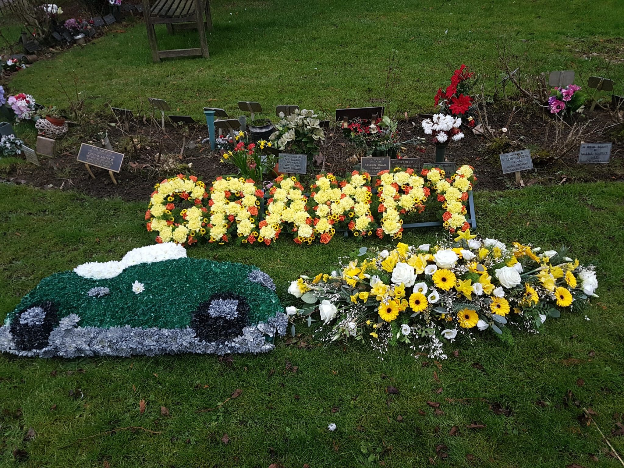 Floral funeral tributes on grass: yellow flowers spelling "GRAMPY," a green car made from flowers, and a yellow and white flower arrangement, with plaques and other flowers in the background.