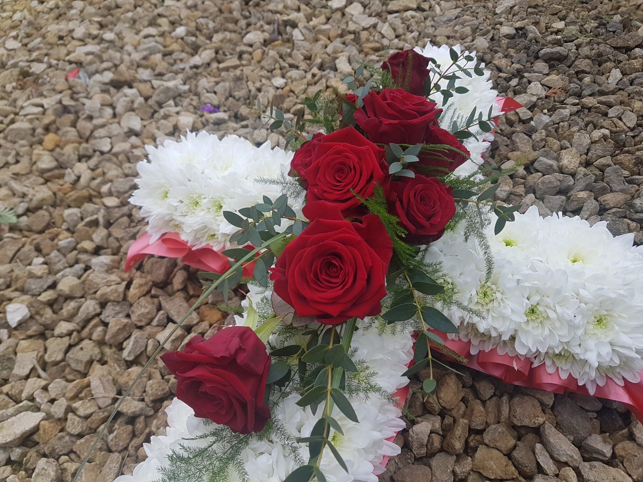 A floral arrangement shaped like a cross lies on gravel, featuring red roses and greenery on a base of white flowers with some pink ribbon visible underneath.