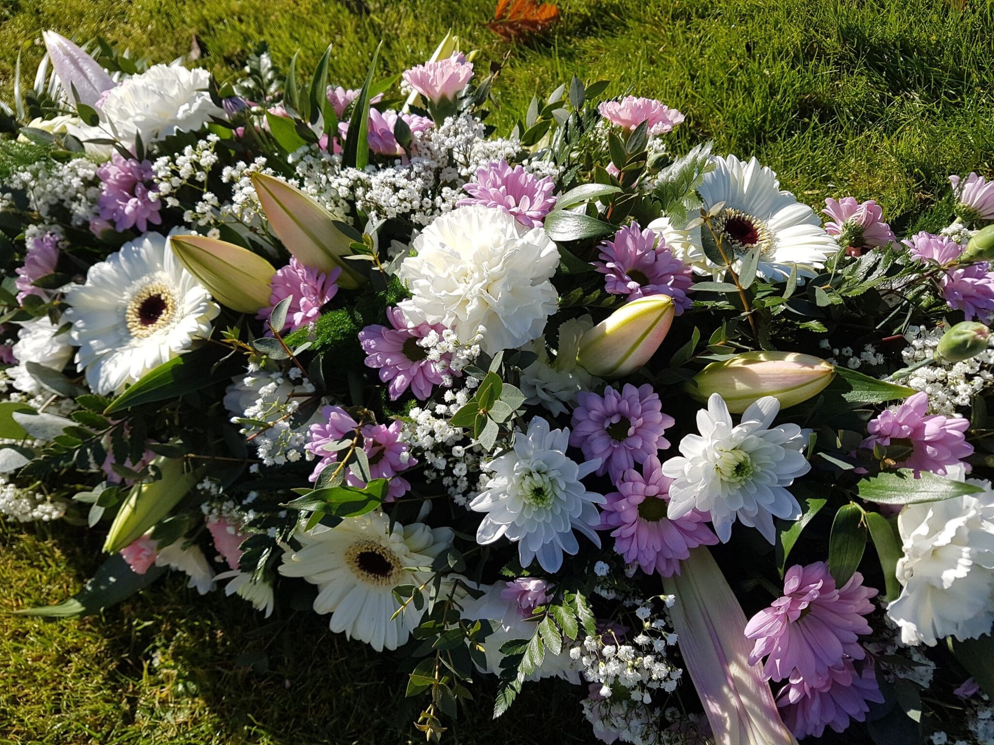 A floral arrangement of white and light purple flowers, including daisies, lilies, and baby's breath, is laid on green grass, possibly as a tribute or at a memorial site.
