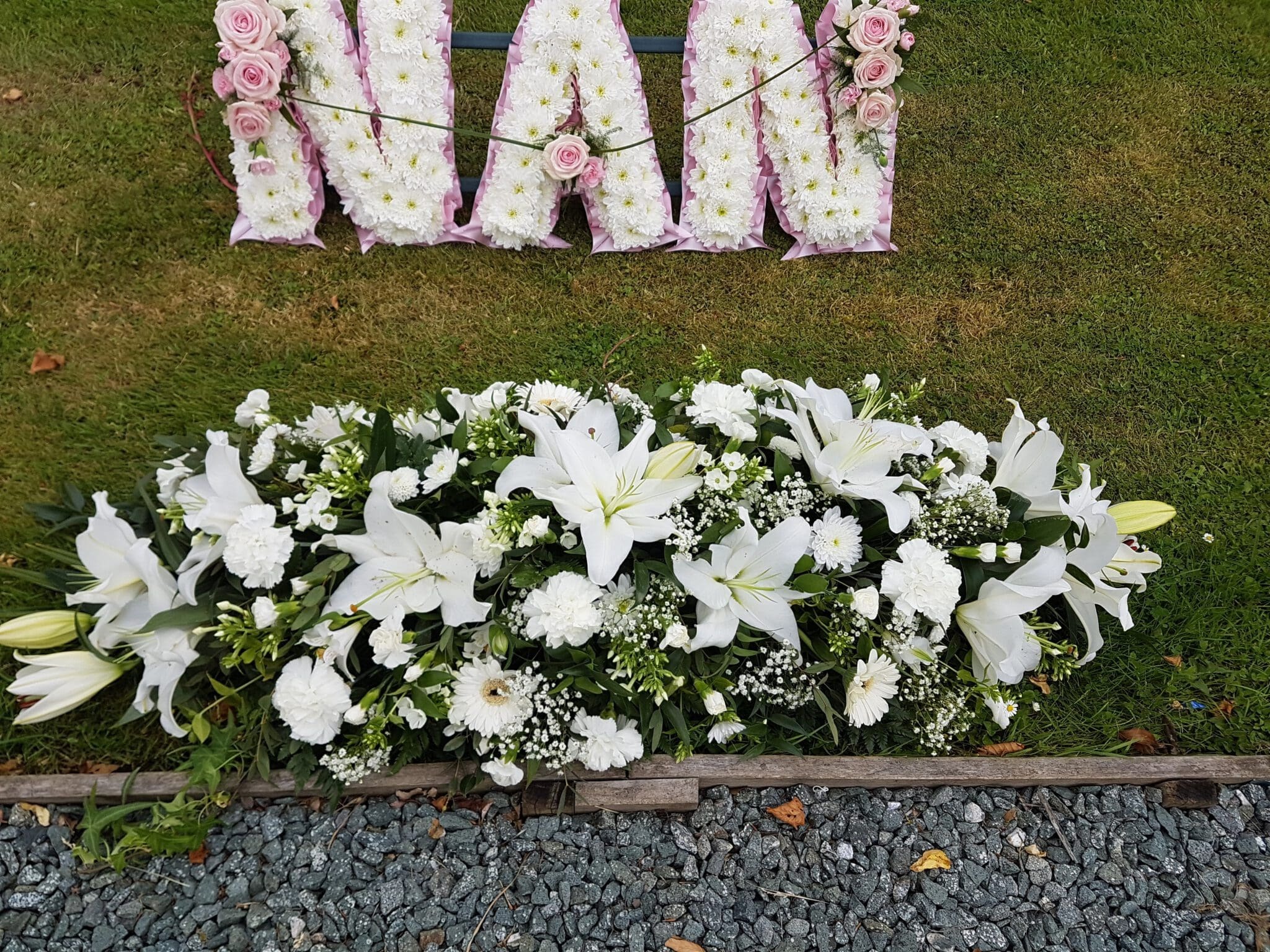 A floral funeral arrangement with white lilies and carnations on grass, below a flower tribute spelling “NAN” in white and pink flowers. Gravel is in the foreground.