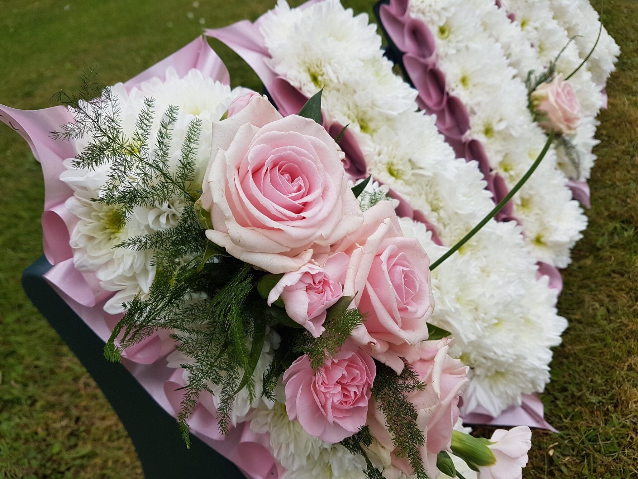A close-up of a floral arrangement featuring pink roses, white chrysanthemums, and greenery on a grassy surface, likely part of a funeral wreath or tribute display.