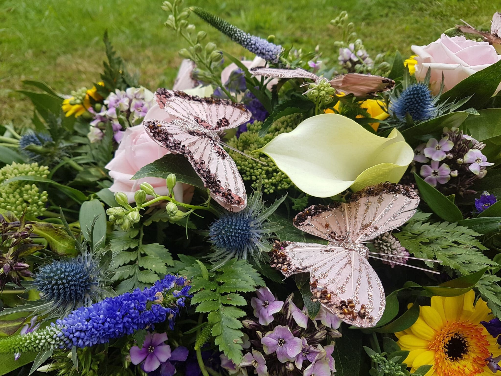 A colorful bouquet of assorted flowers, including yellow lilies, purple and blue blossoms, pink roses, and ferns, adorned with two decorative pink butterflies, set against a grassy background.