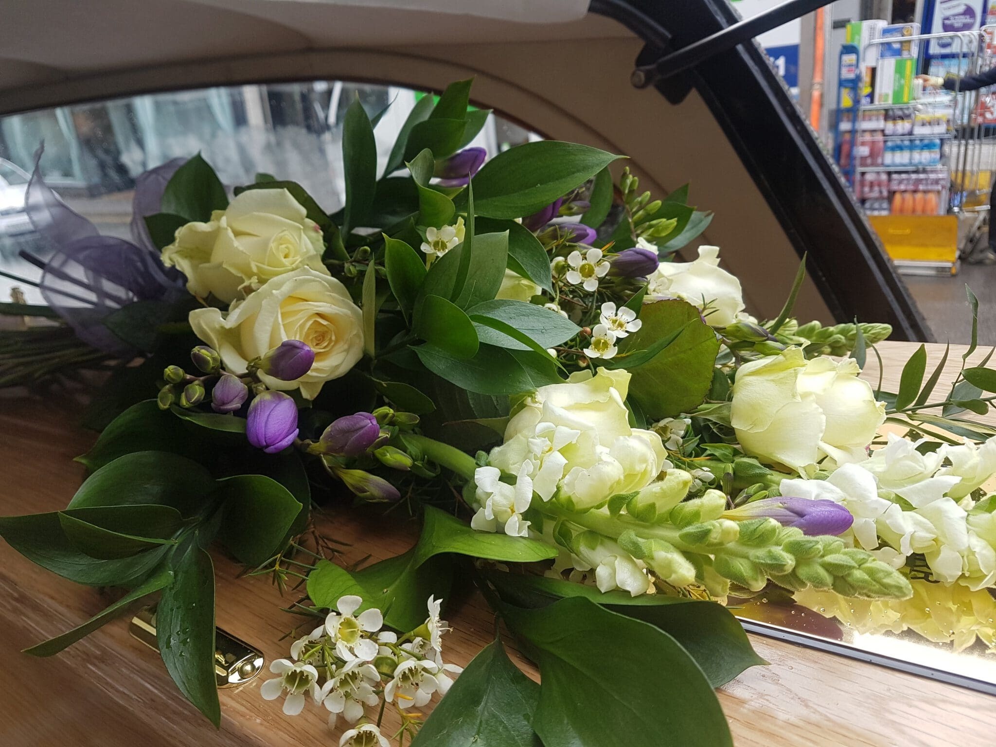 A floral arrangement of white roses, purple flowers, and greenery rests on a wooden surface, possibly inside a vehicle, with a shop visible through the window in the background.