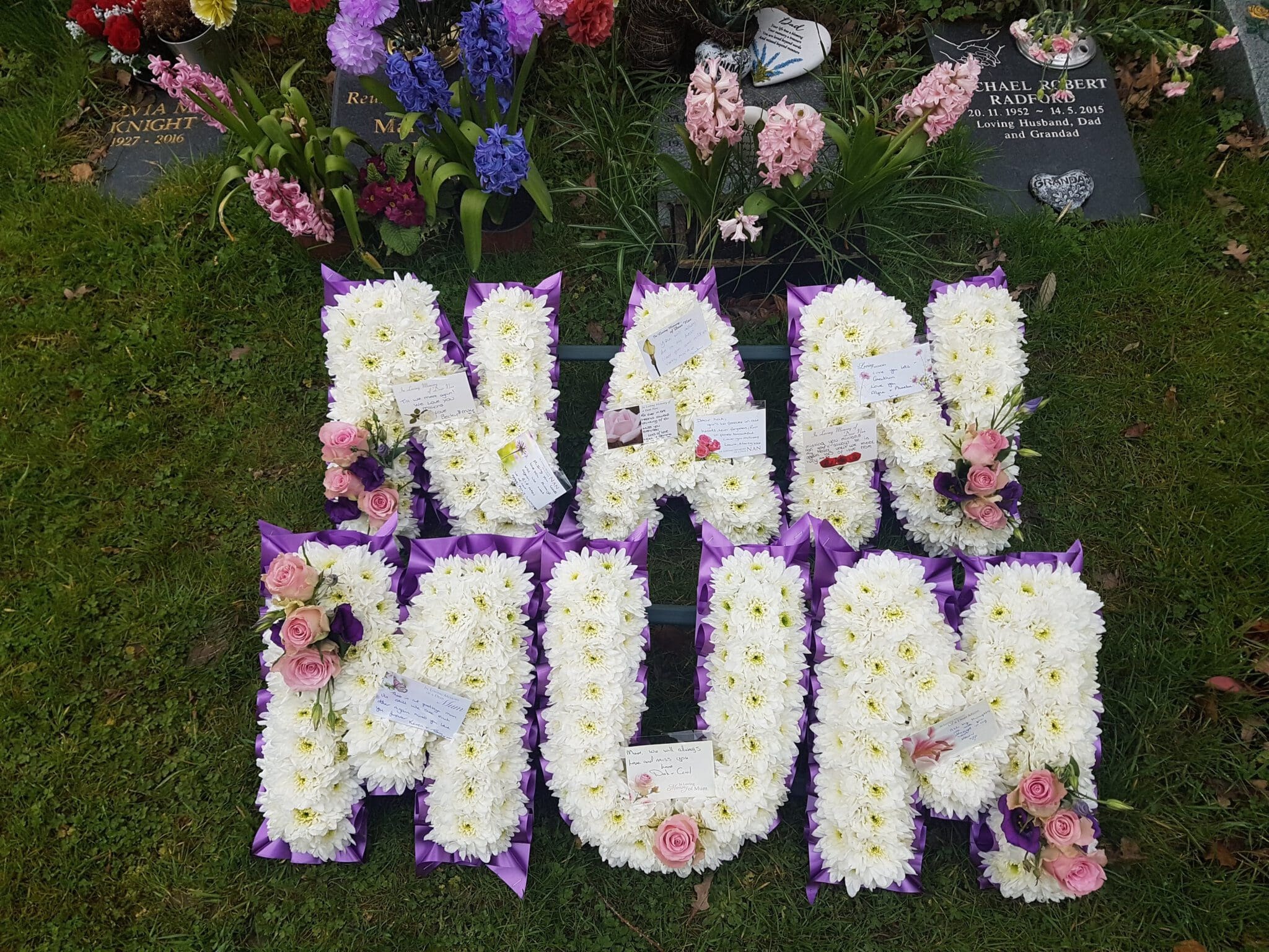 Floral tributes on grass arranged to spell "NAN" and "MUM" in large white and purple flowers, accented with pink roses, at a cemetery with headstones and other flower arrangements in the background.