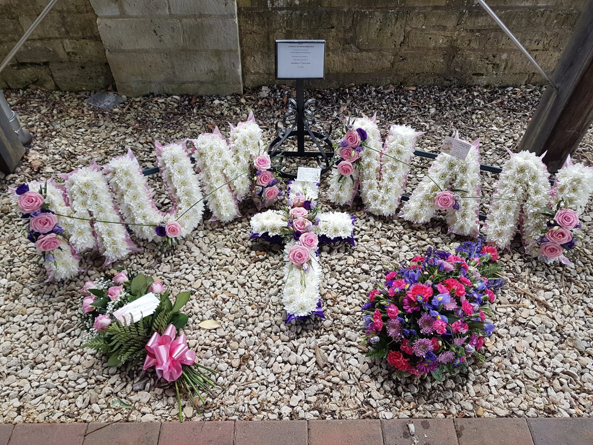 Floral funeral tributes arranged on gravel: large white letters with pink roses spelling "HUMNAN," a cross made of white flowers, and two other flower arrangements, one circular and one with green leaves and pink bow.