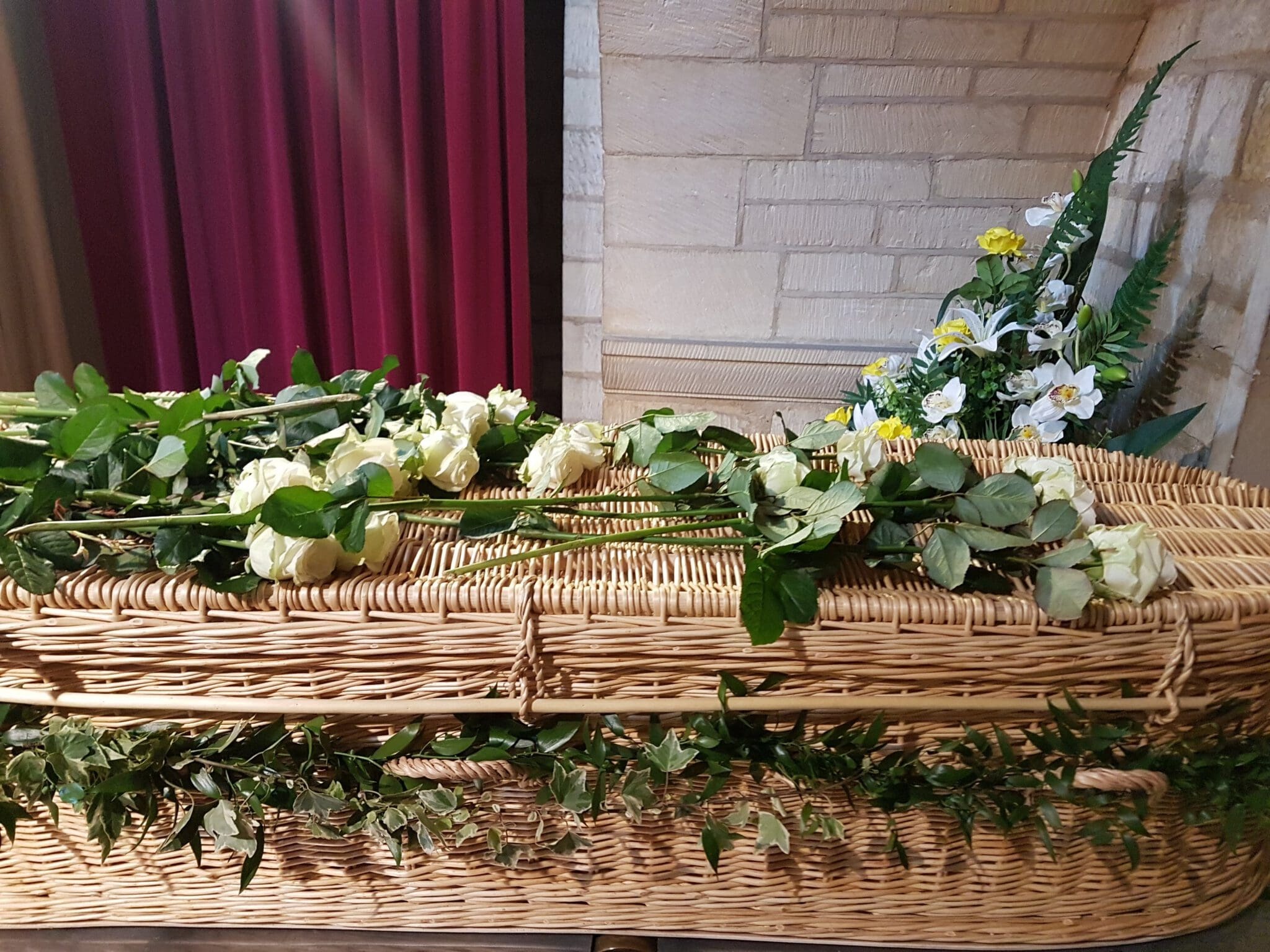 A wicker coffin adorned with white roses and greenery sits in a room with red curtains and a stone wall. Floral arrangements are visible in the background.