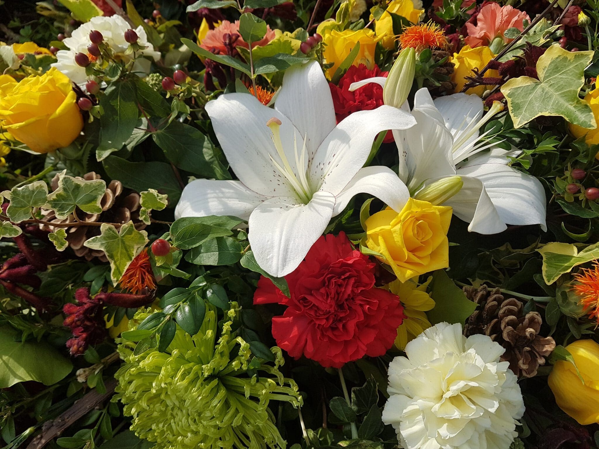 A vibrant floral arrangement featuring white lilies, yellow roses, red and white carnations, green chrysanthemums, ivy leaves, pinecones, and orange flowers.