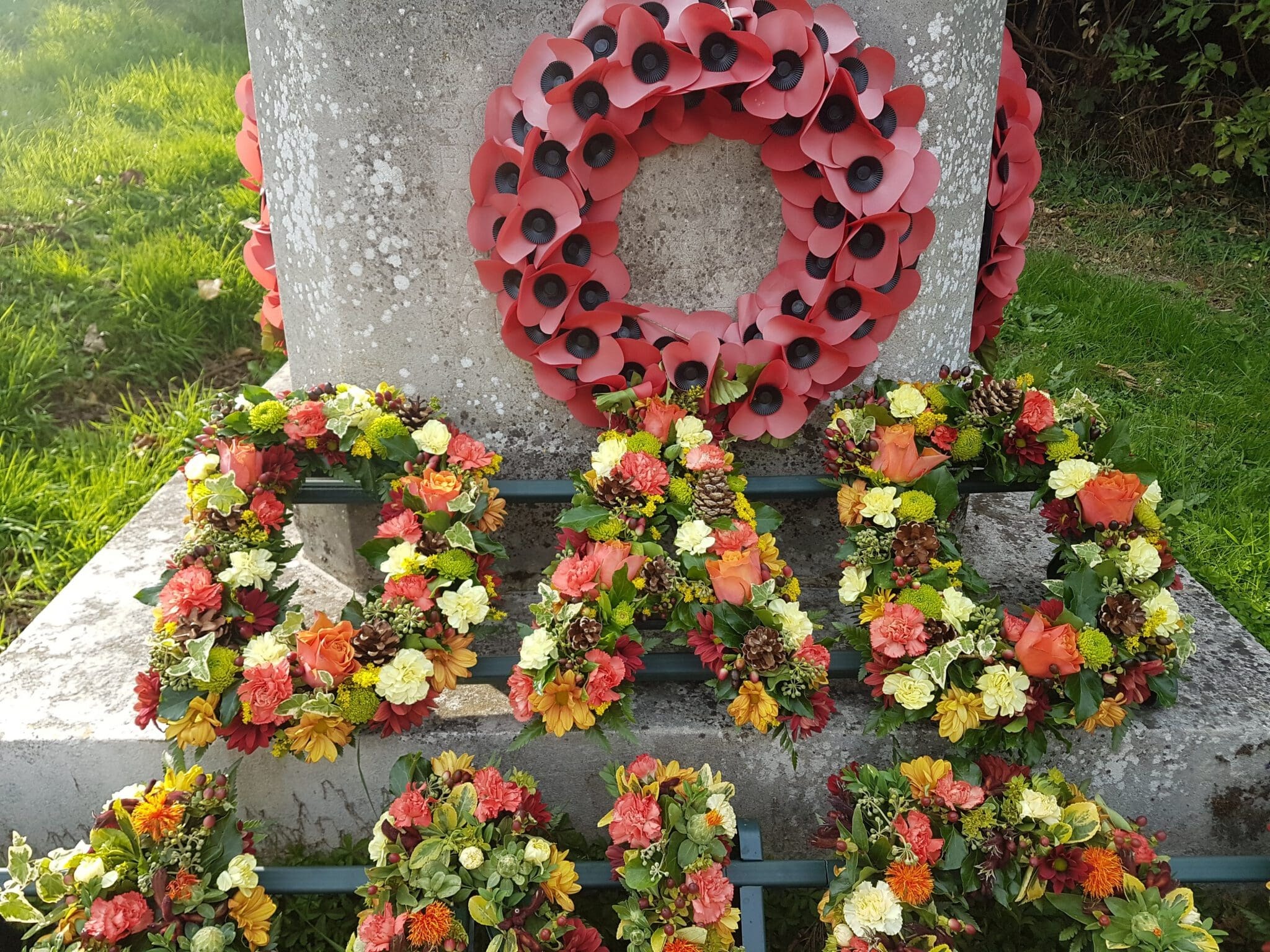 A memorial stone with wreaths of red poppies and colorful flowers arranged to form the letters "CAD" on a sunny grassy area.