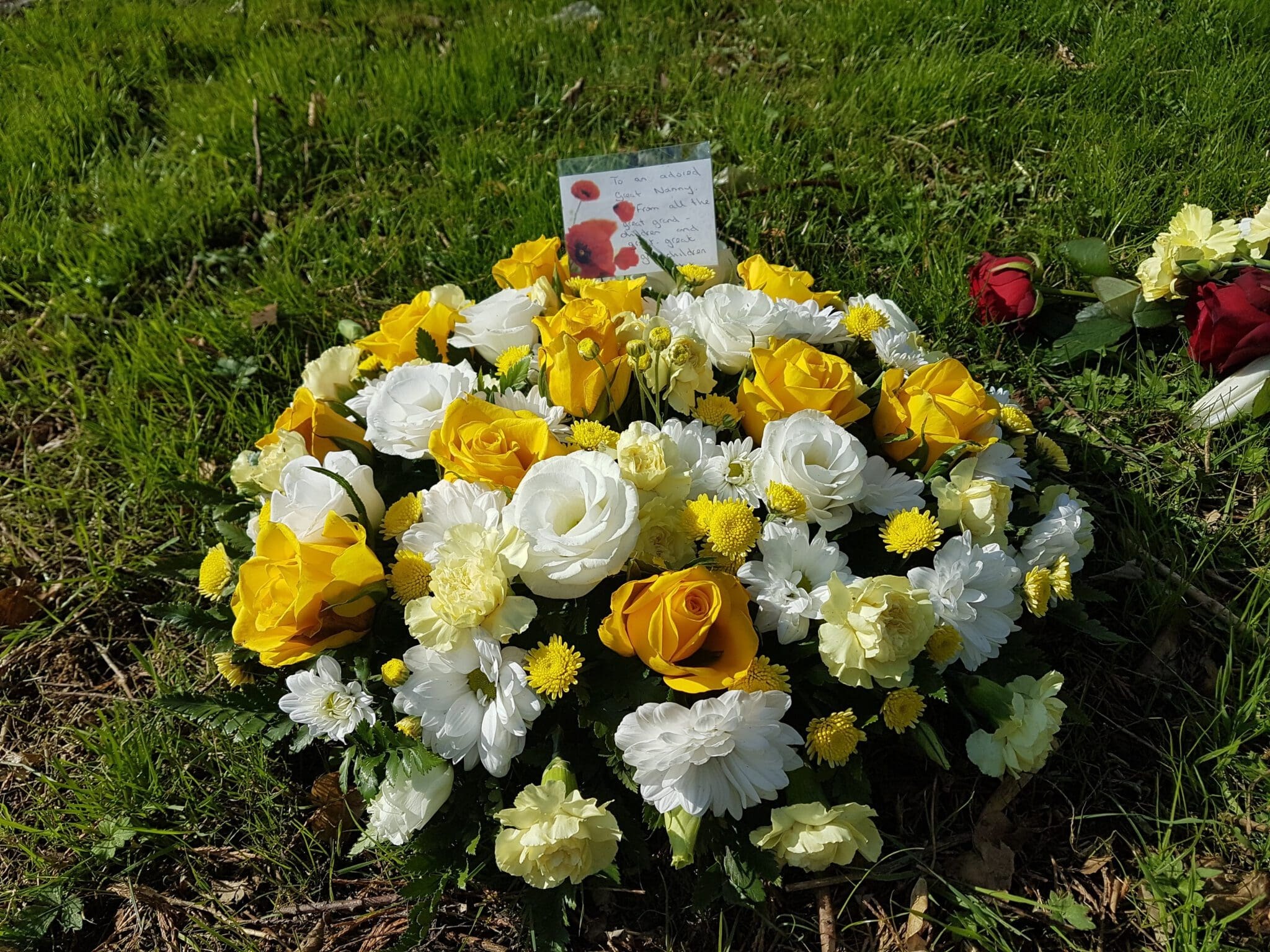 A round floral arrangement of yellow and white flowers, including roses and daisies, sits on green grass with a card and a single red rose nearby.