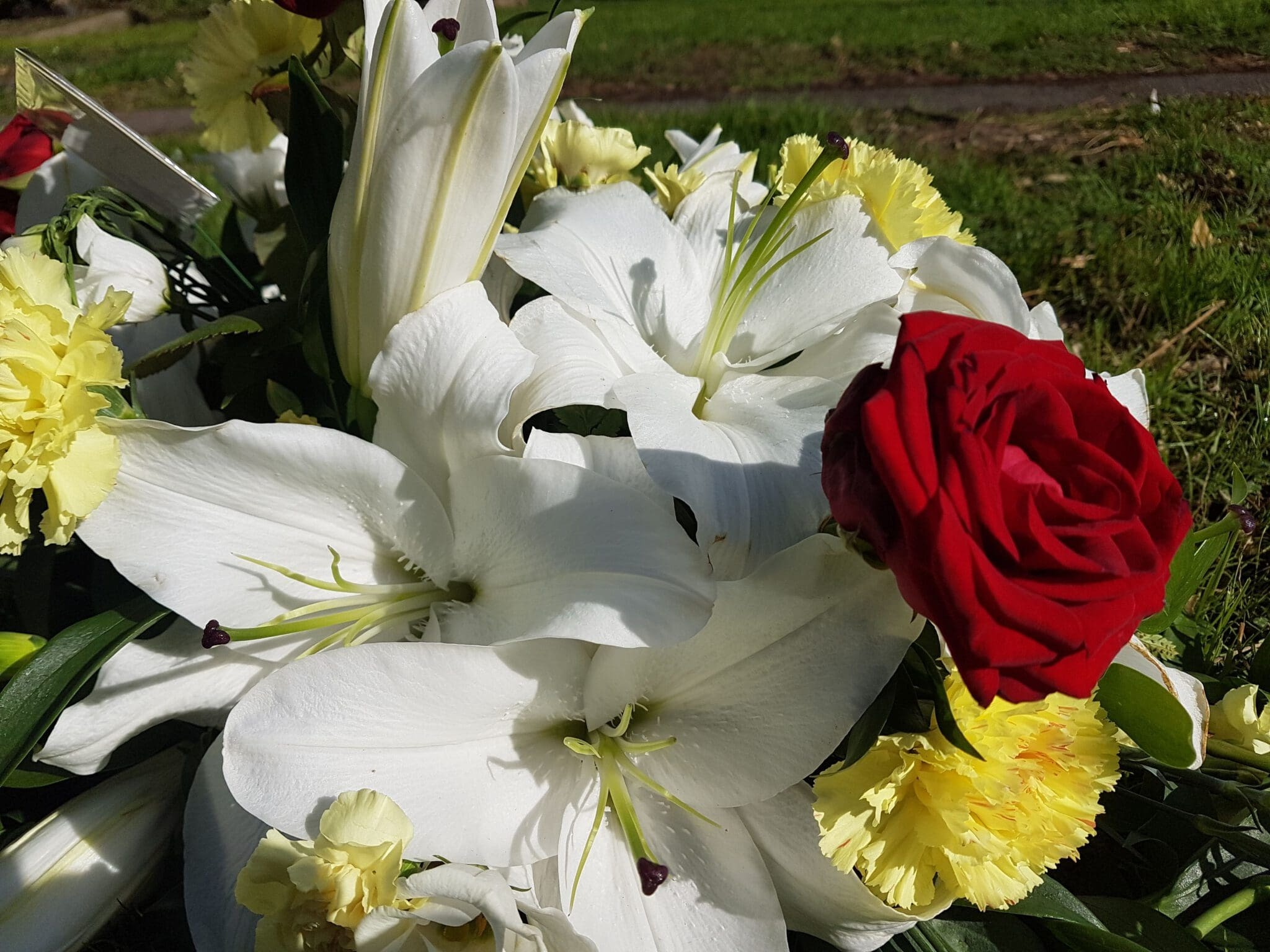 A close-up of a floral arrangement featuring white lilies, yellow carnations, and a single red rose, set outdoors on green grass in sunlight.