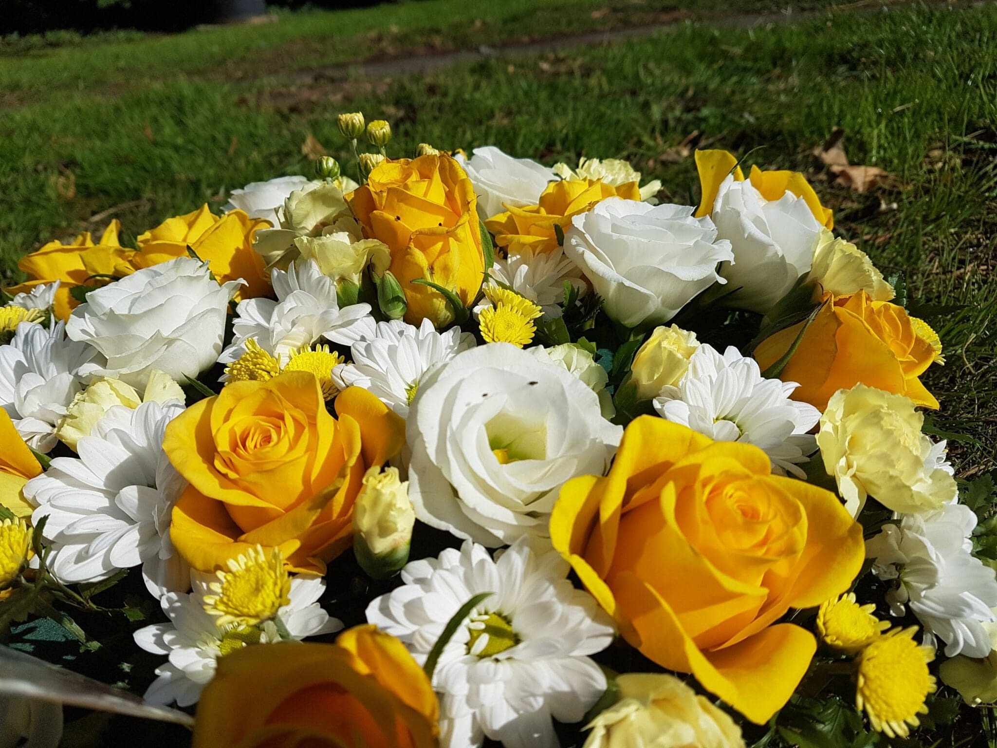 A close-up of a bouquet with yellow and white roses, white daisies, and small yellow flowers, set outdoors on a background of green grass.