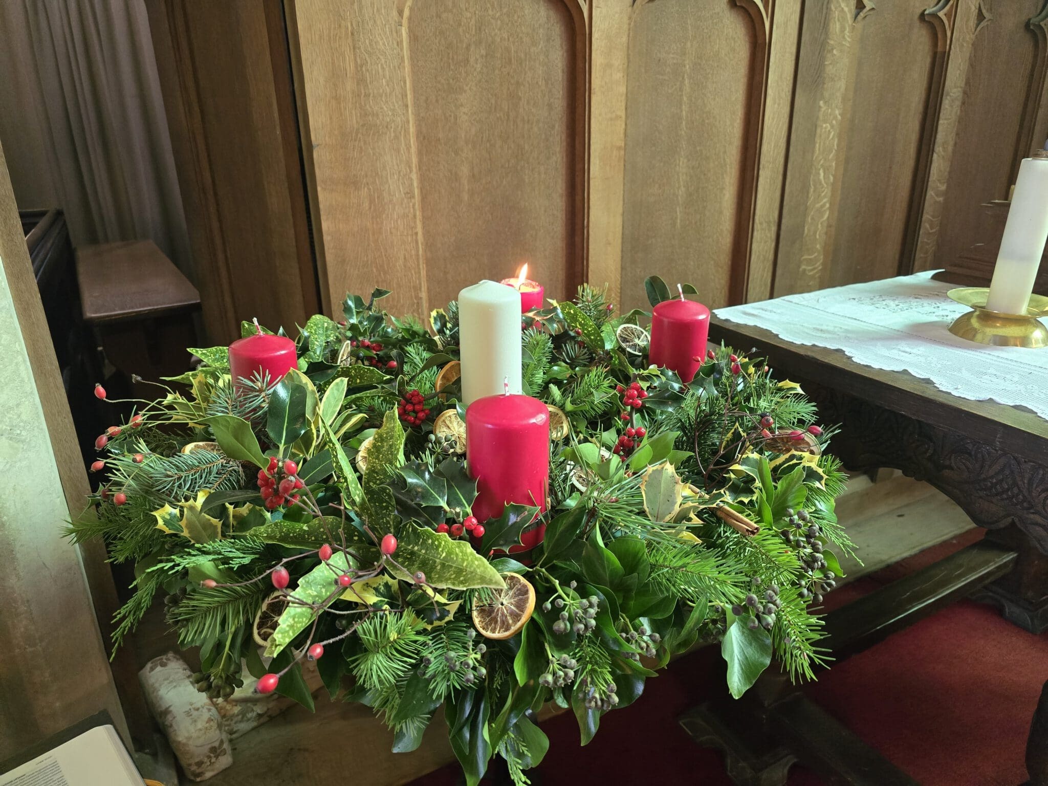 An Advent wreath with four red candles and one white candle is decorated with greenery, berries, and dried orange slices. One red candle is lit, and the wreath is placed on a wooden table inside a church.