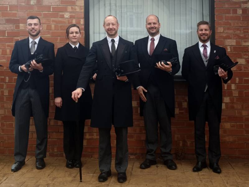 Five people dressed formally in black coats and striped trousers stand in a row in front of a brick wall, each holding a black top hat. The group includes four men and one woman, all smiling at the camera.