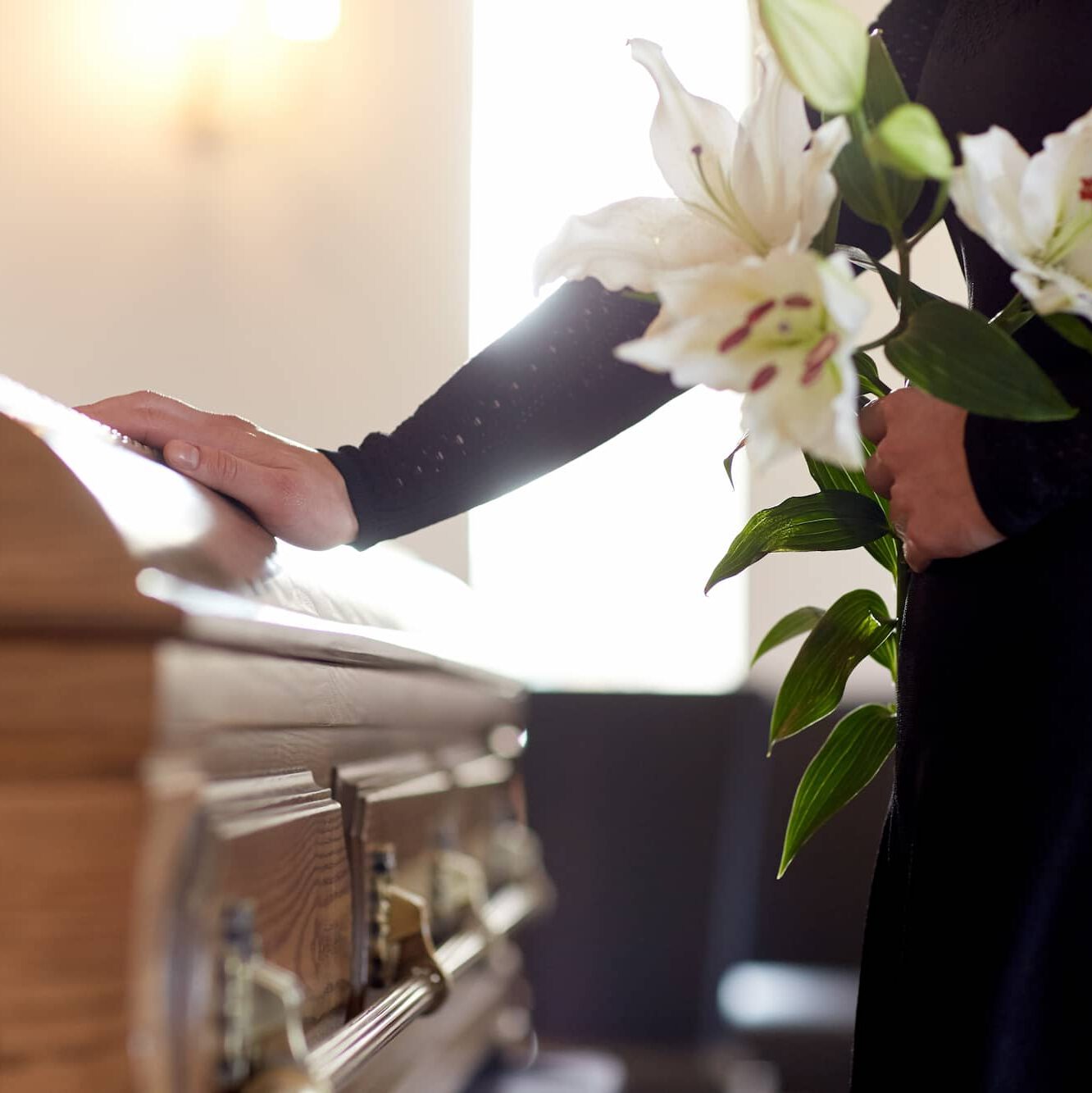 A person in black stands beside a wooden casket, resting one hand on it and holding white lilies in the other, in a softly lit room, suggesting a funeral or memorial service.