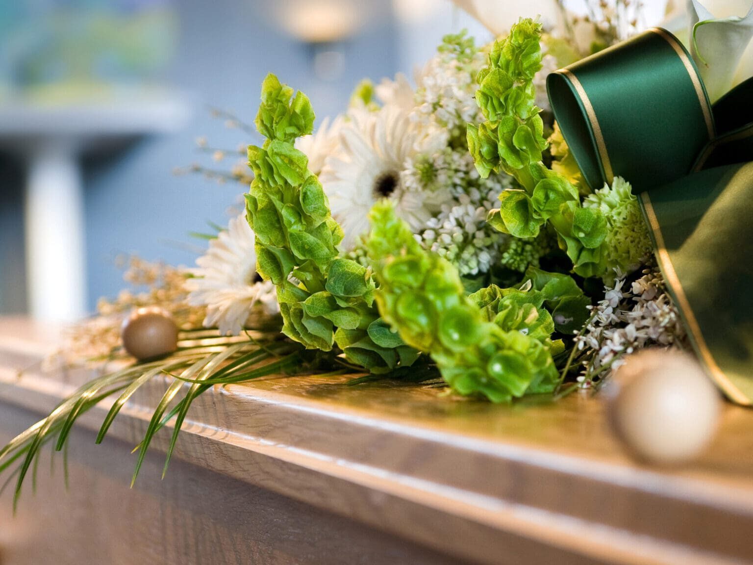A close-up of a wooden casket adorned with a bouquet of white flowers, green leaves, and a dark green ribbon, symbolizing a funeral setting.