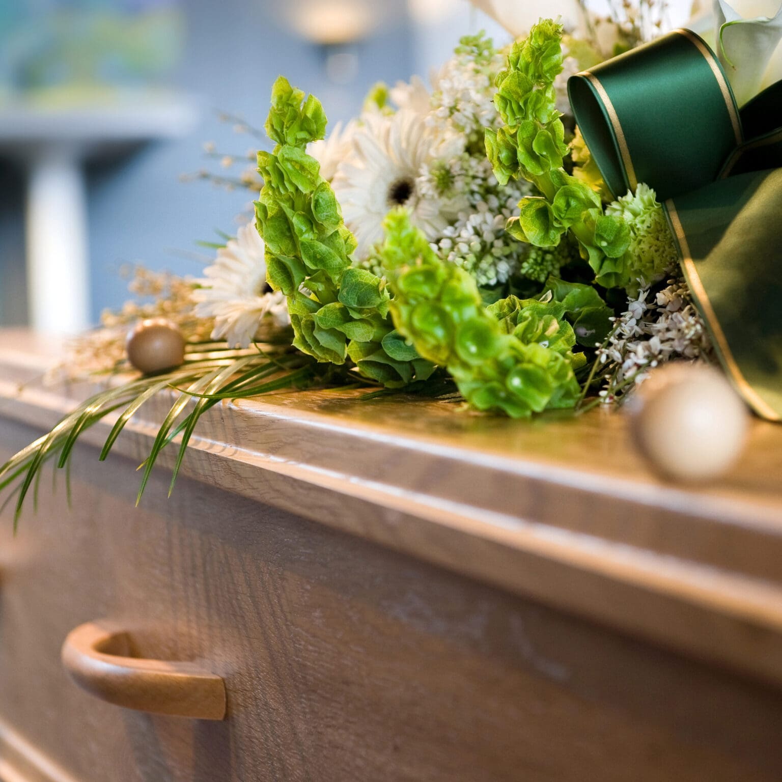 A close-up of a wooden coffin with a flower arrangement of white blooms and green leaves on top, with blurred background details.