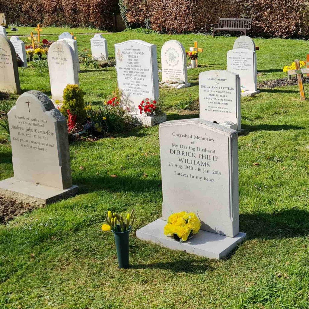 A gravestone in a sunny cemetery, inscribed with "Derrick Philip Williams" and adorned with a vase of yellow flowers. Other headstones and colorful flower arrangements are visible in the background, along with a bench.