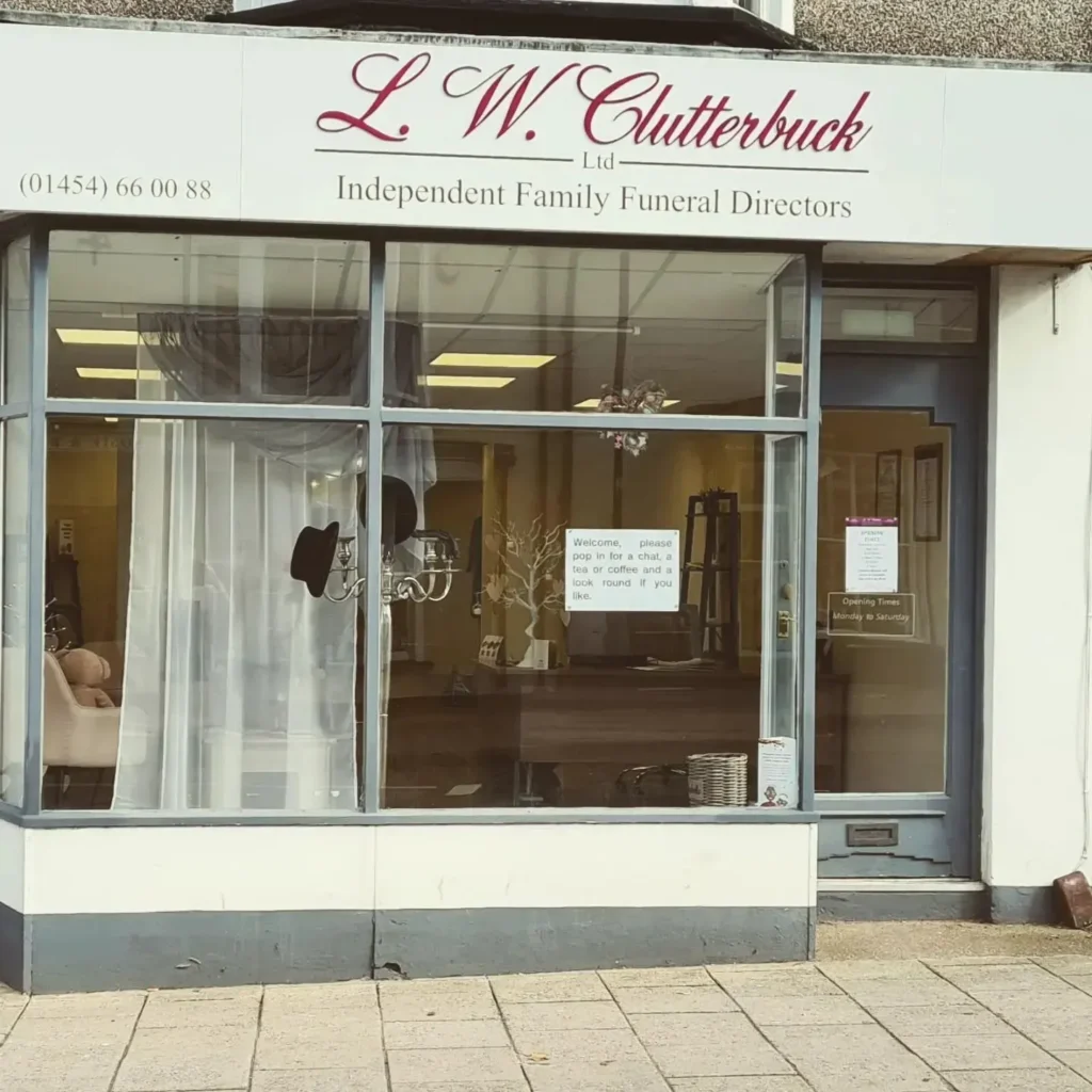 Shopfront of L.W. Clutterbuck Ltd, an independent family funeral directors, with large windows, white curtains, and a sign displaying their phone number above the entrance.