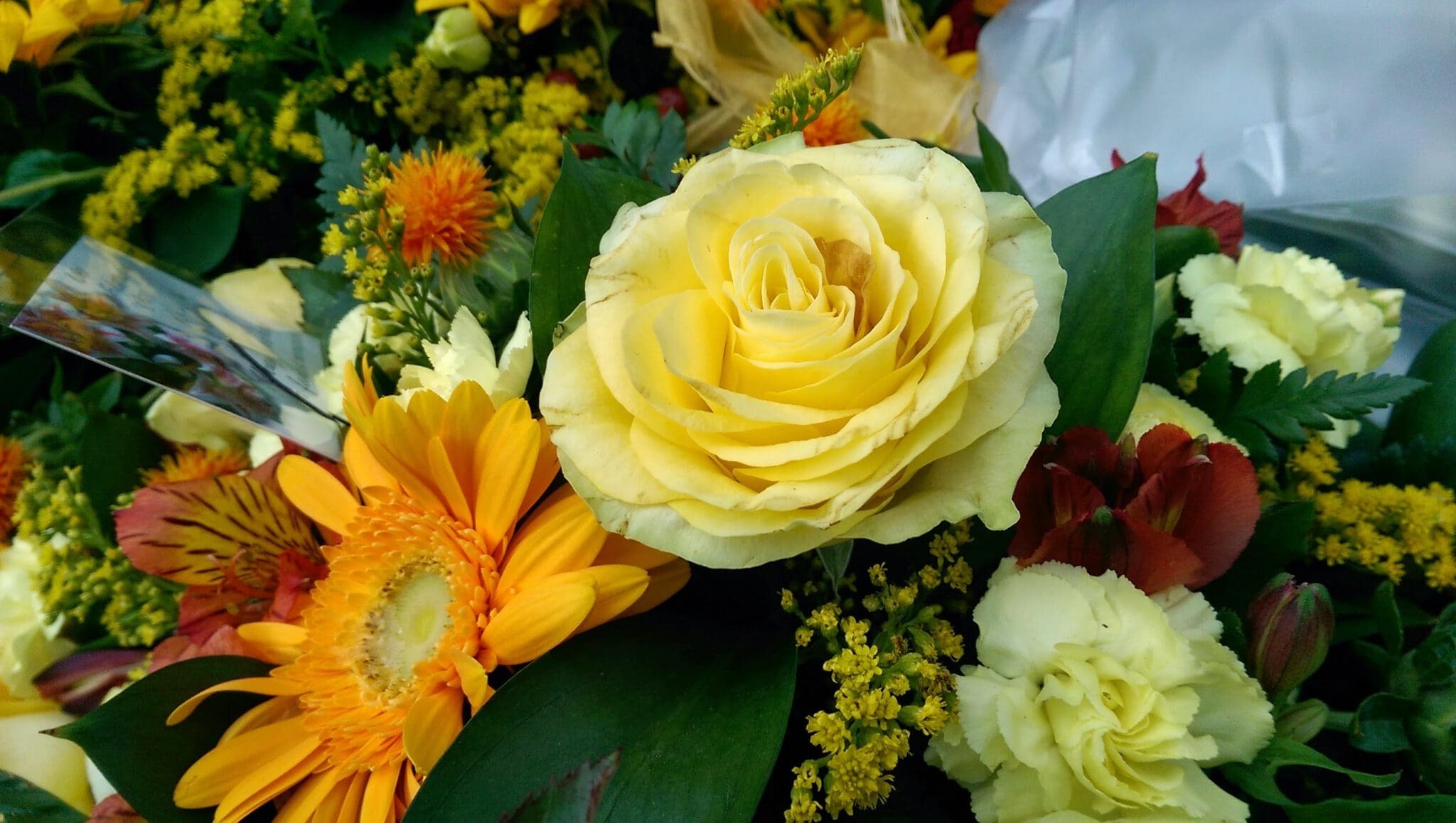 A vibrant bouquet featuring a yellow rose, yellow gerbera daisy, small orange and red flowers, green leaves, and sprigs of yellow filler flowers. The arrangement is bright and cheerful.
