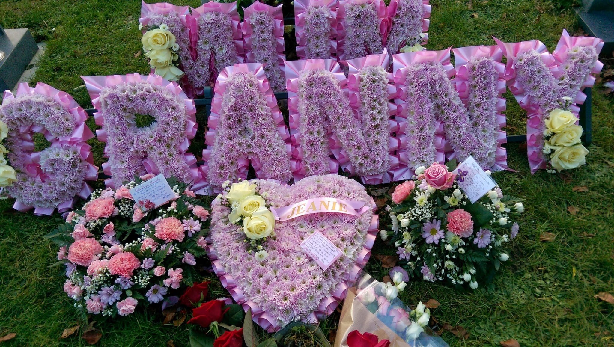 Pink floral funeral tribute spelling "GRANNY" with white and pink flowers, heart-shaped arrangement labeled "JEANNE," and various flower bouquets placed on green grass.