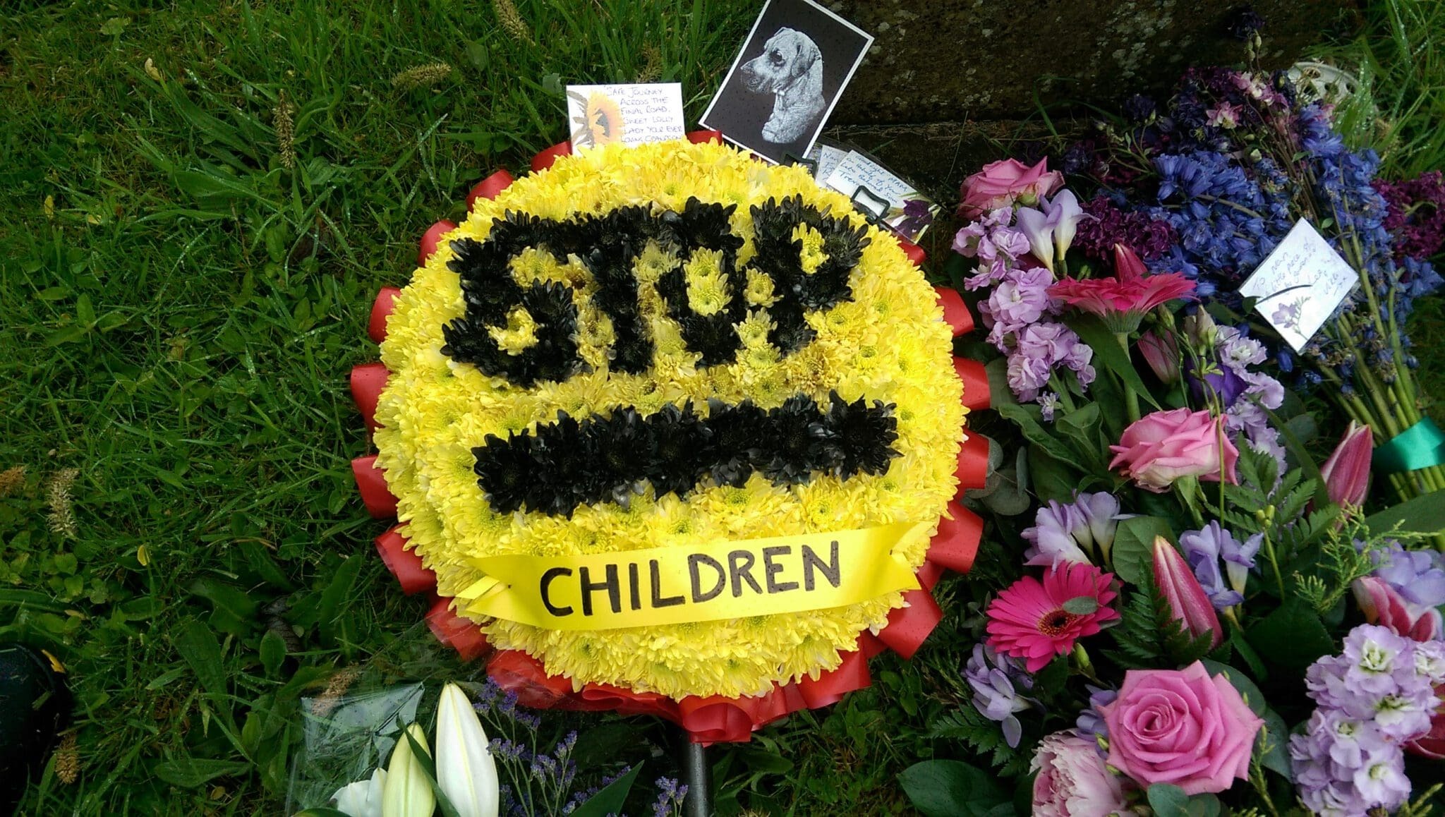 A yellow and red floral wreath with "STOP" in black flowers and "CHILDREN" on a yellow ribbon, surrounded by purple, pink, and white flower arrangements on green grass.