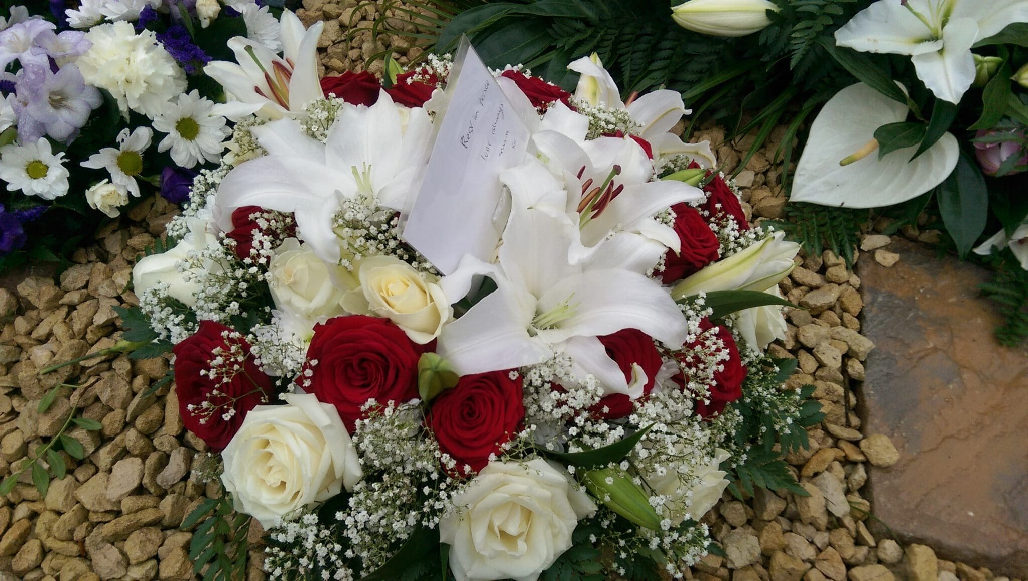 A funeral wreath with white lilies, red and white roses, baby's breath, and a card, placed on a bed of small stones with other floral arrangements nearby.