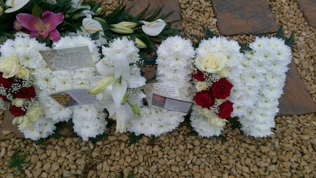 A floral tribute spelling "MUM" in white flowers with clusters of red and white roses, lilies, and handwritten cards, placed on gravel and surrounded by more flowers.