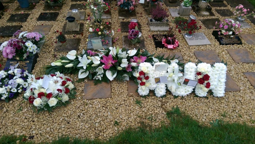 A graveside display with floral arrangements, including lilies and roses, and a prominent tribute spelling "DAD" in white flowers, set among gravestones and gravel.