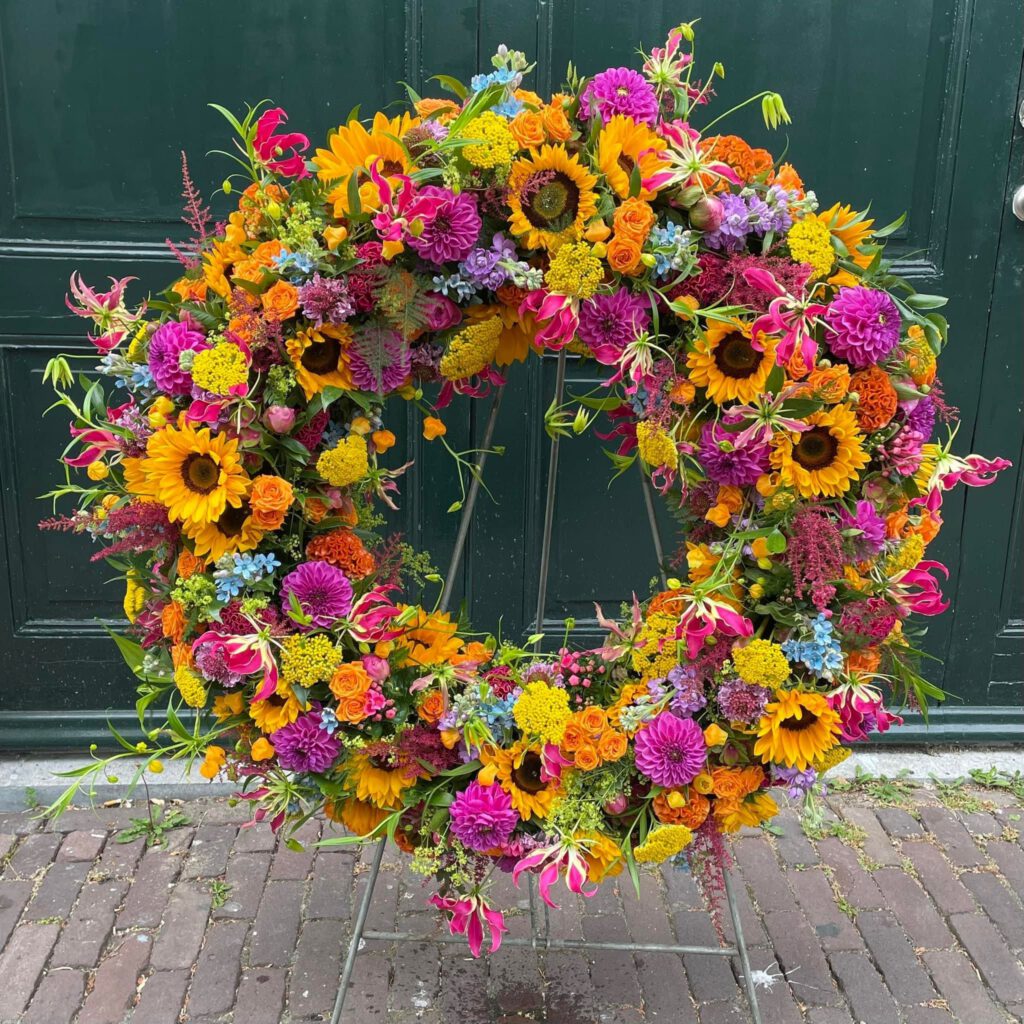 A large, vibrant floral wreath with sunflowers, dahlias, roses, and other colorful flowers in shades of yellow, pink, orange, and purple, displayed on a stand in front of a dark green door.
