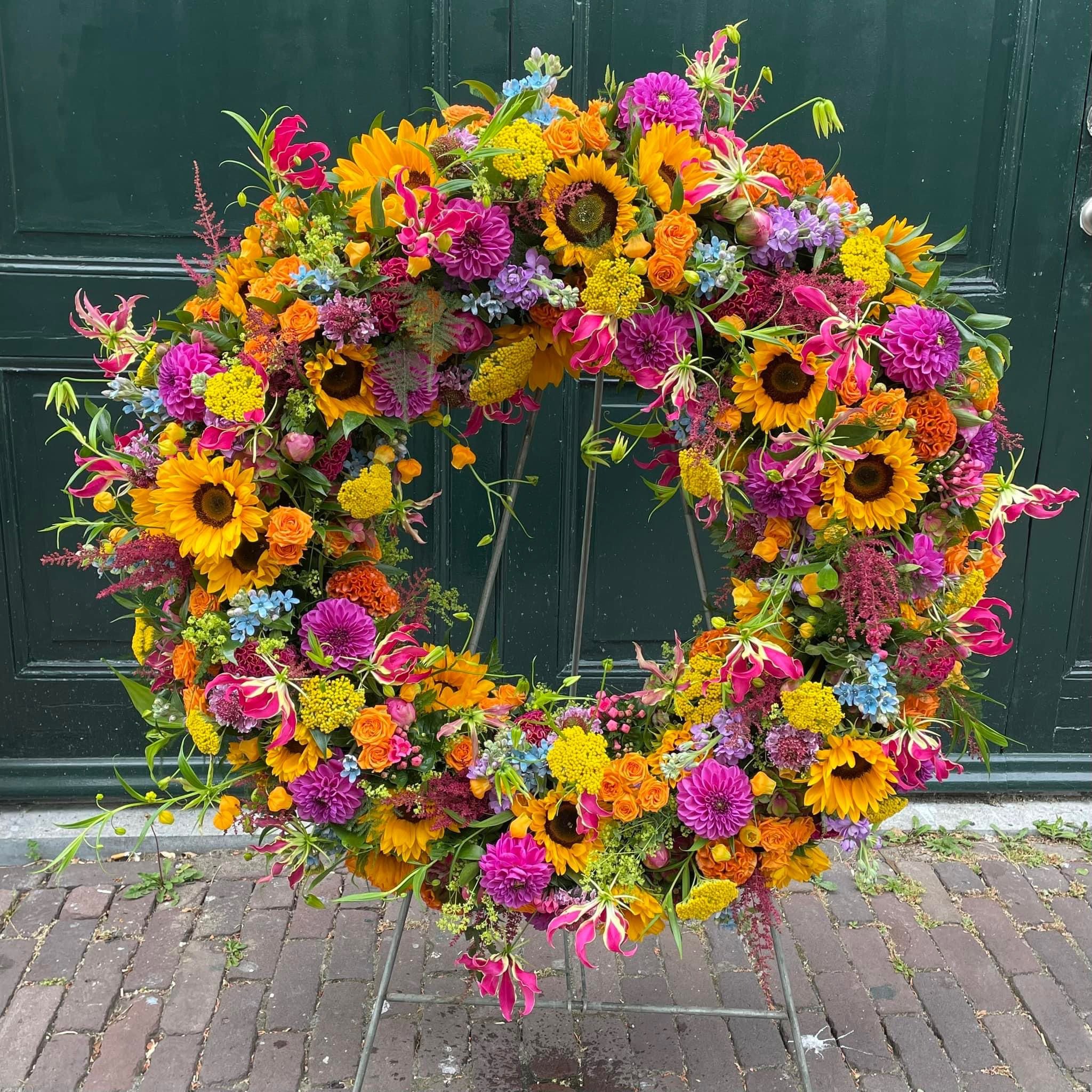 A large, vibrant floral wreath with sunflowers, dahlias, roses, and other colorful flowers in shades of yellow, pink, orange, and purple, displayed on a stand in front of a dark green door.