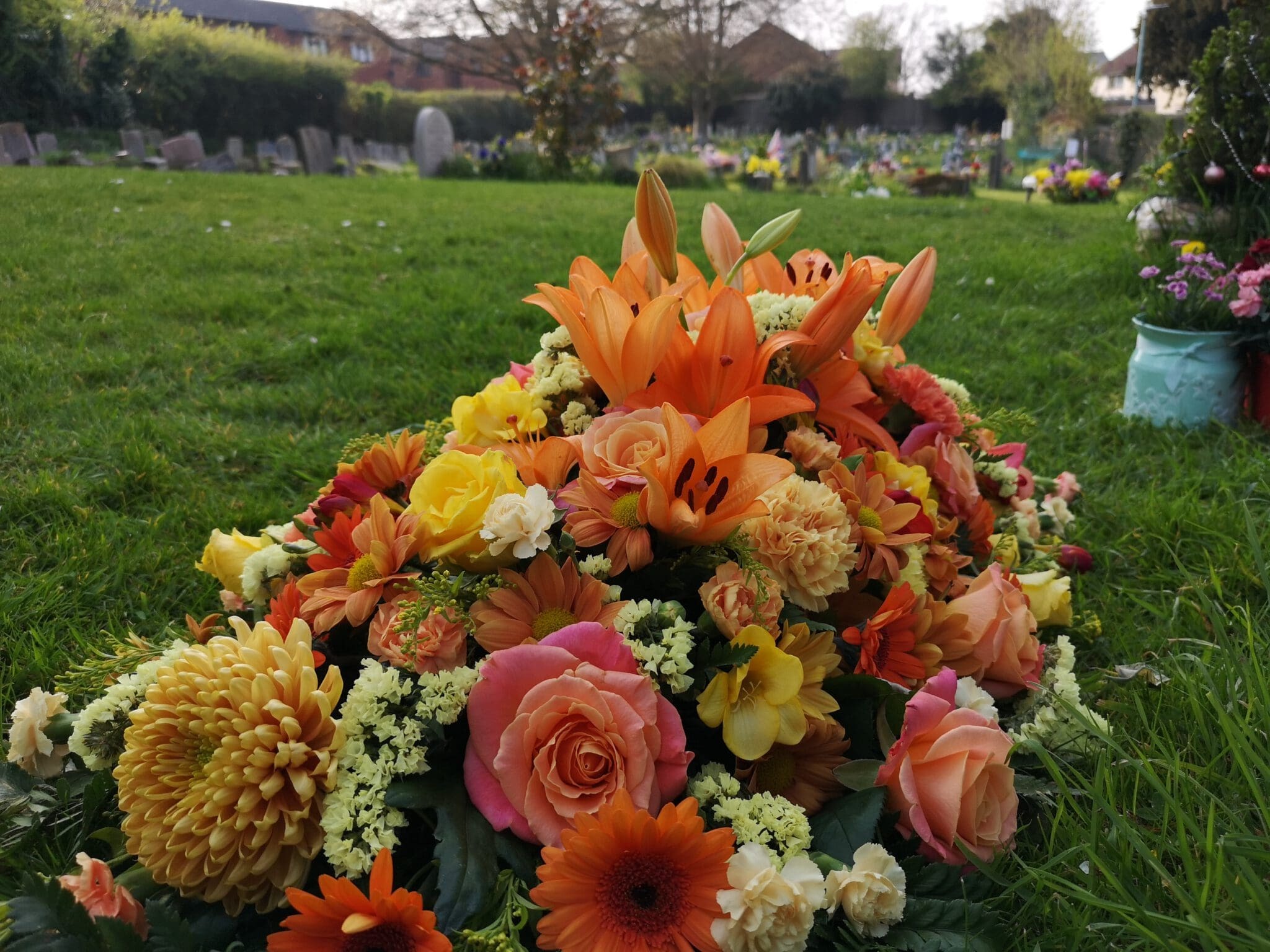 A colorful floral arrangement with lilies, roses, and chrysanthemums rests on green grass in a cemetery, with headstones and more flowers visible in the background.