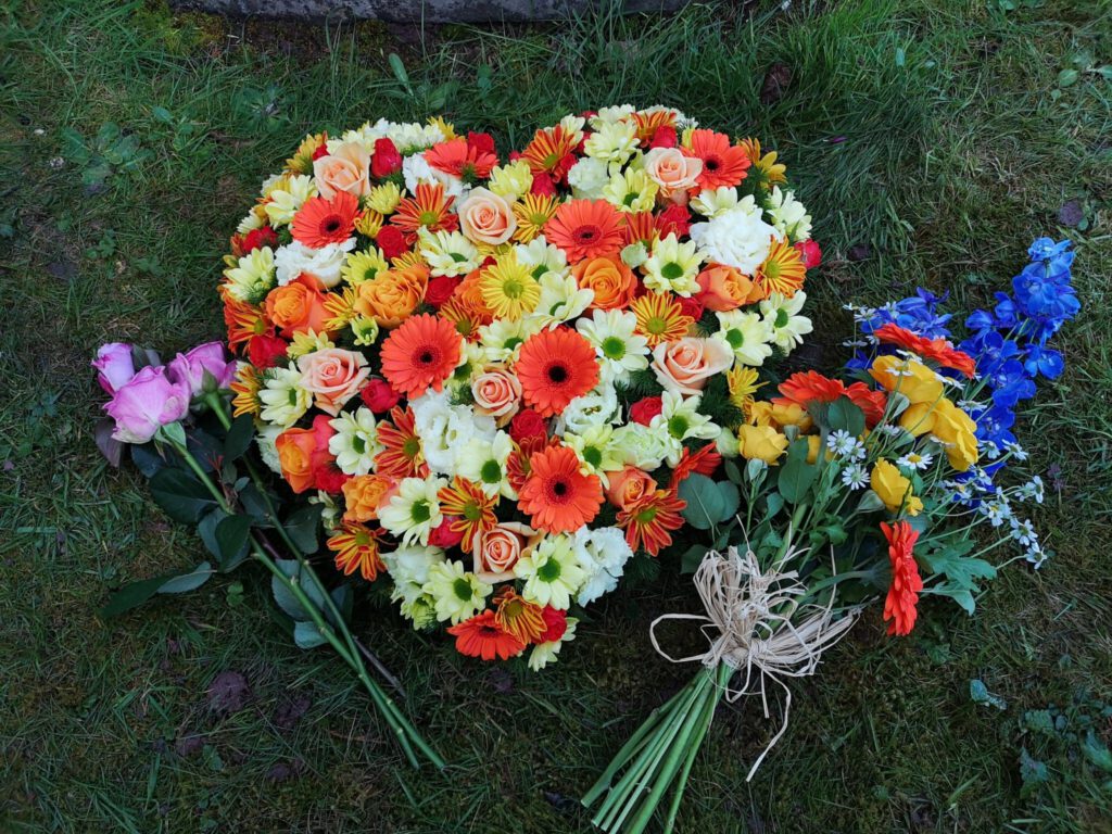 A heart-shaped bouquet of orange, yellow, and white flowers lies on grass, with two smaller arrangements: one of pink roses and another with blue, yellow, and orange flowers tied with twine.