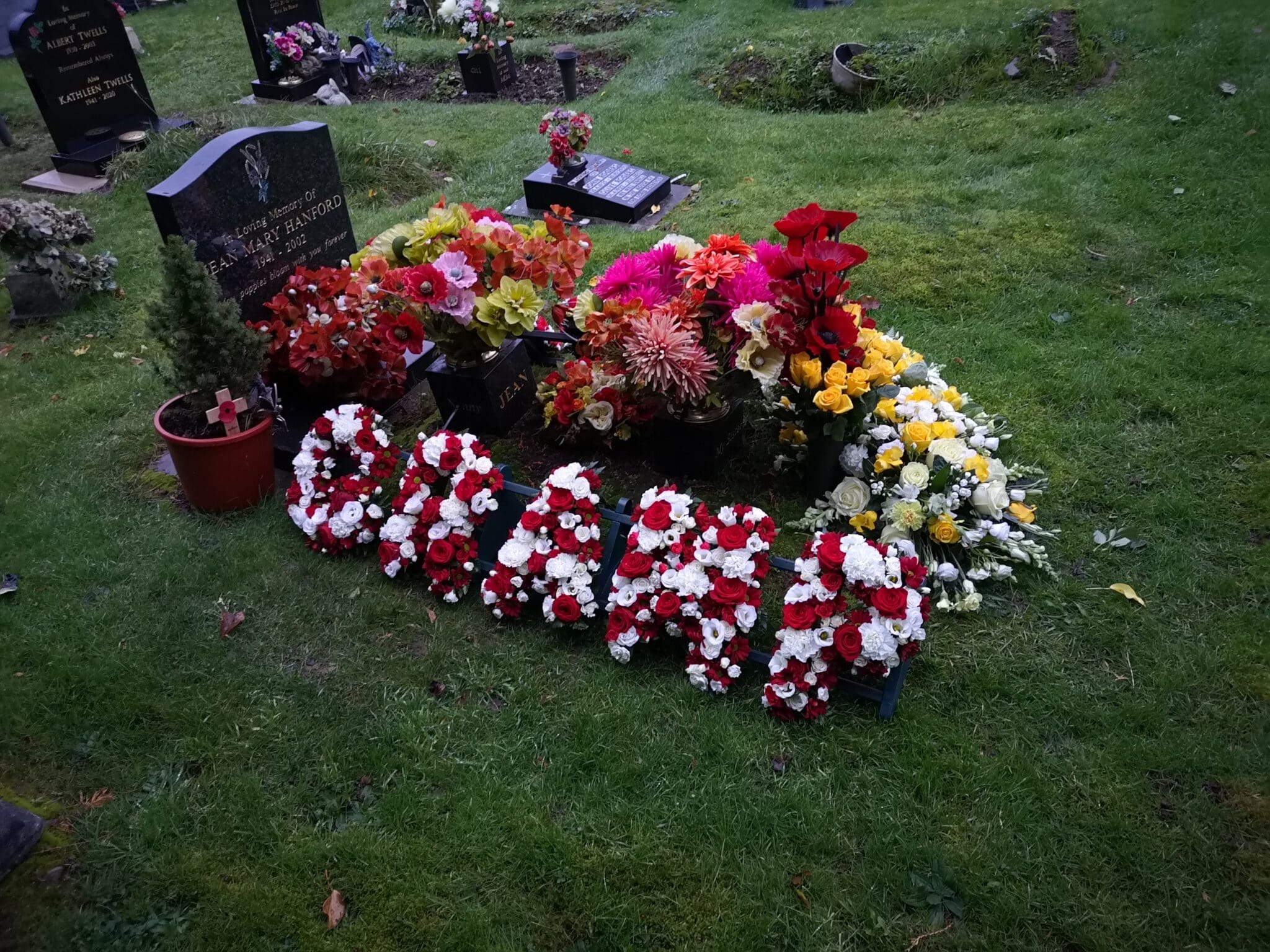 A grave decorated with colorful flower arrangements and wreaths, including red, white, yellow, and pink flowers, with floral letters spelling out "GRANDAD" resting on the grass in front of the headstone.