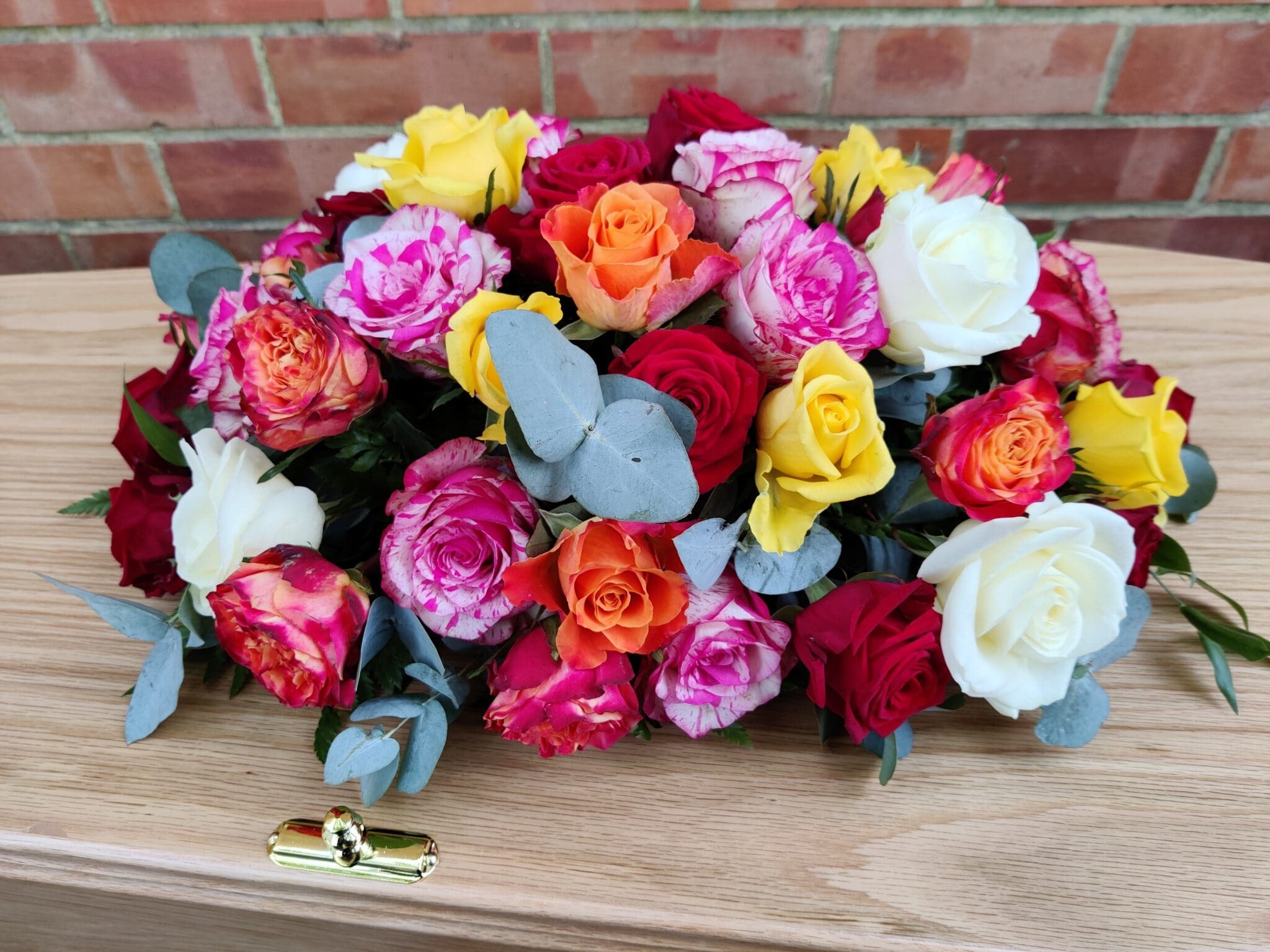 A colorful floral arrangement of roses in red, pink, yellow, orange, and white with green leaves sits on a wooden coffin, against a brick wall background.