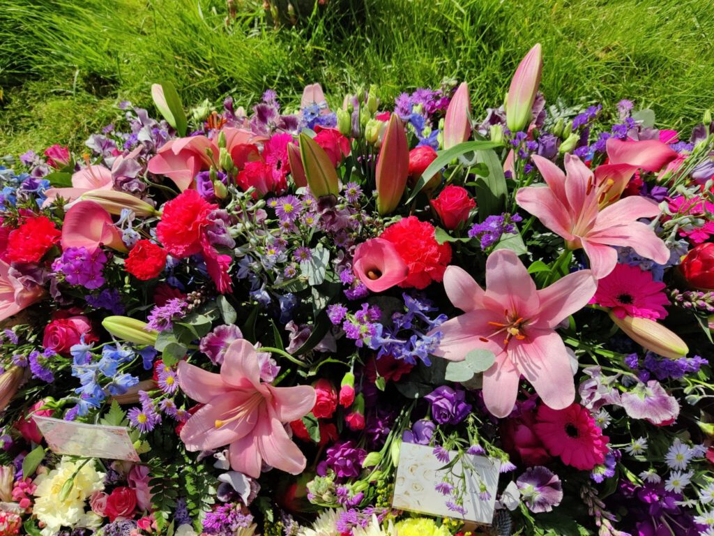 A vibrant arrangement of pink lilies, roses, purple asters, and other colorful flowers is displayed outdoors on green grass, with small cards nestled among the blooms.
