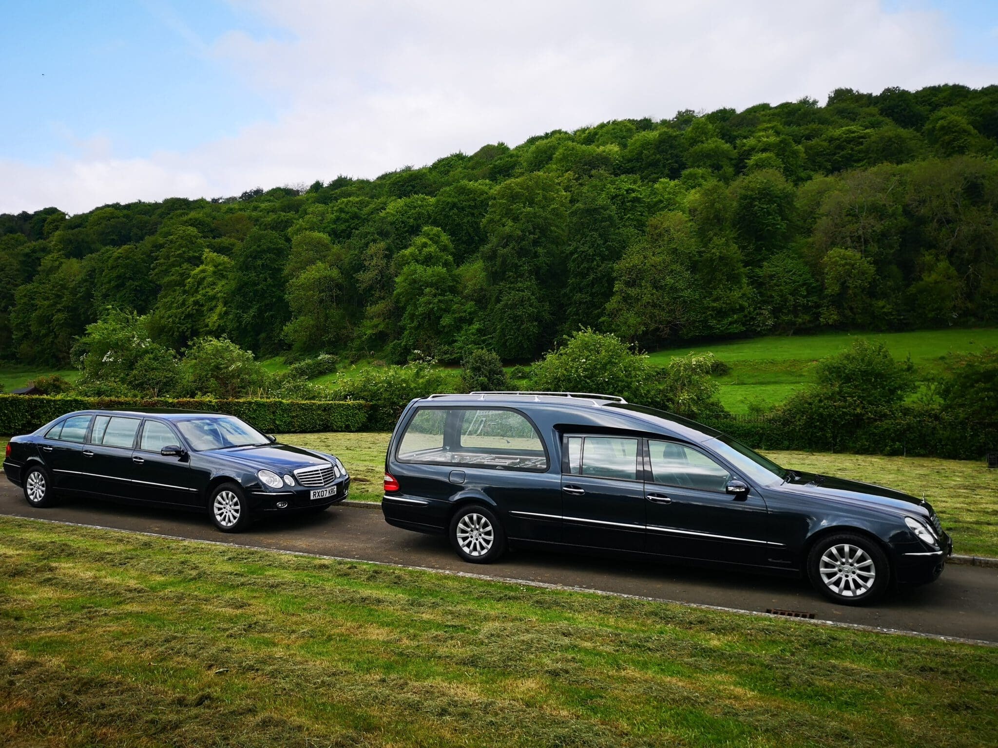 Two black hearses are parked on a paved road beside a grassy area, with a backdrop of dense green trees and a cloudy sky.