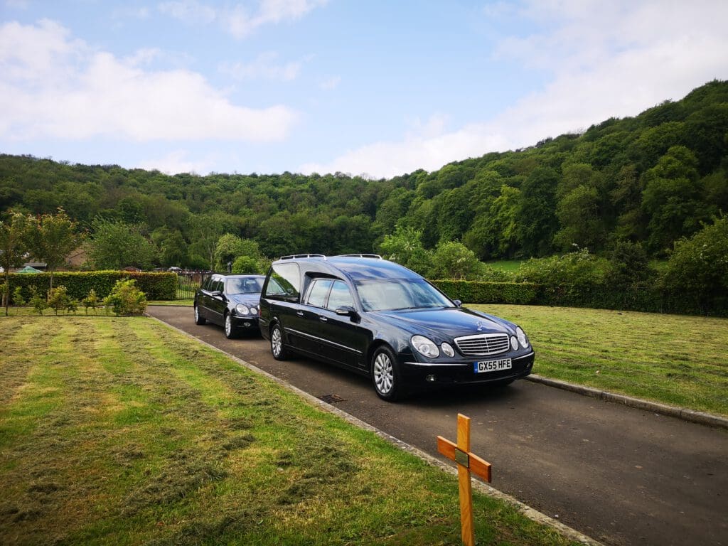 A black hearse and a second car drive along a paved path through a green cemetery, with a wooden cross and freshly cut grass in the foreground, and tree-covered hills in the background under a partly cloudy sky.