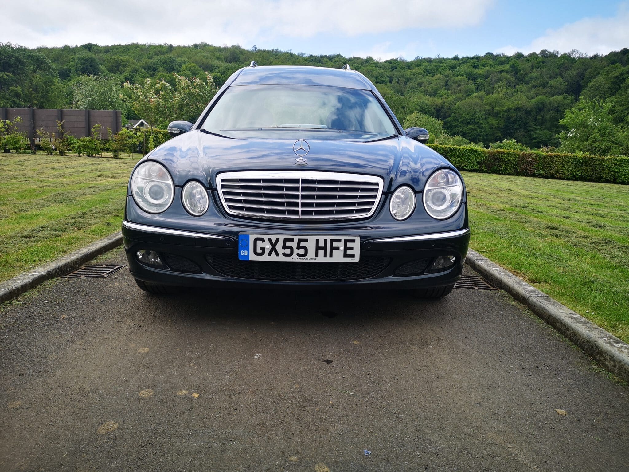 A black Mercedes-Benz car is parked on a paved area with a grassy landscape and trees in the background. The car’s front view shows its headlights, grille, and UK license plate “GX55 HFE.”.