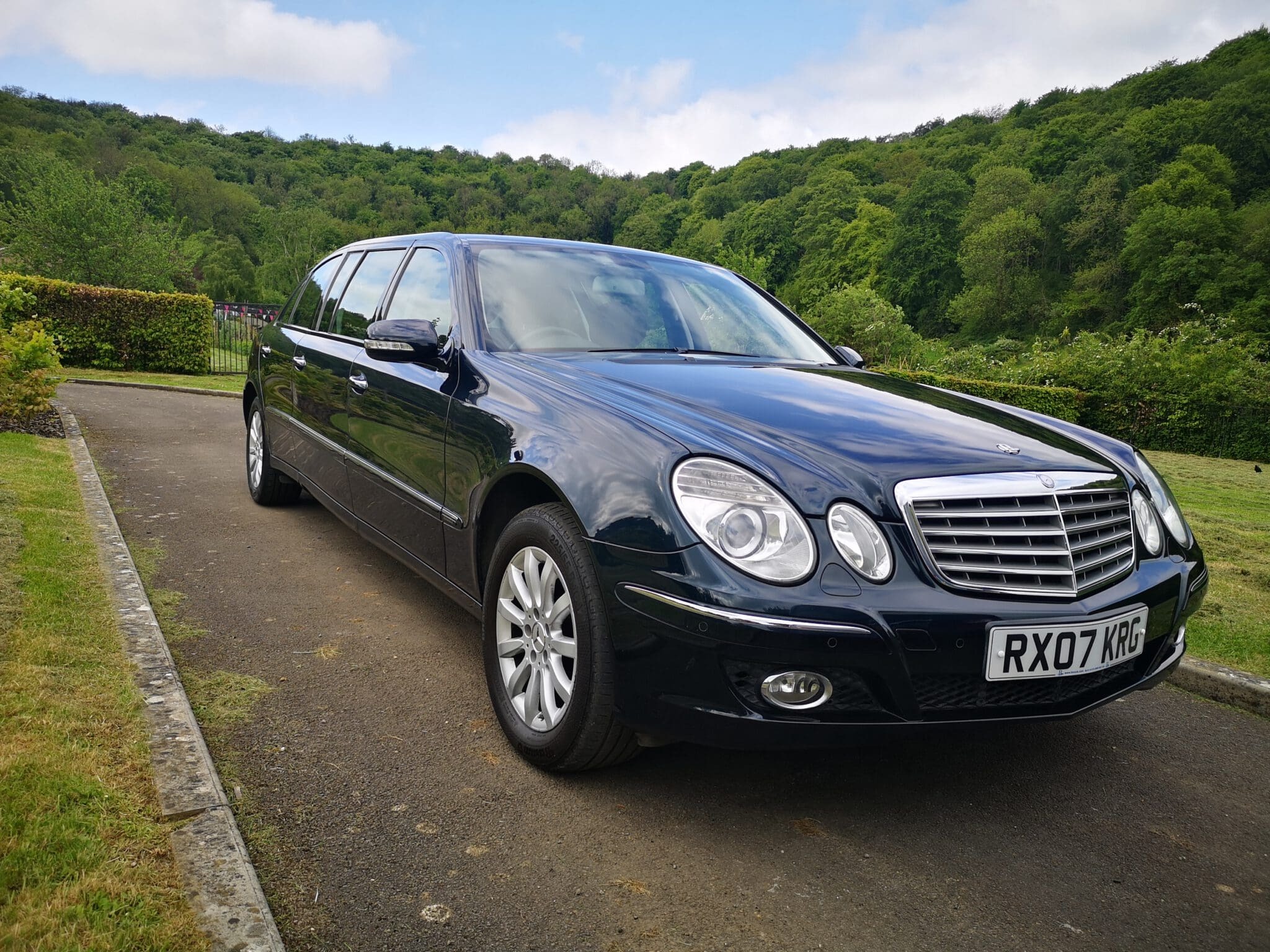 A black limousine with a UK license plate parked on a paved driveway, surrounded by green bushes and trees, under a partly cloudy sky.