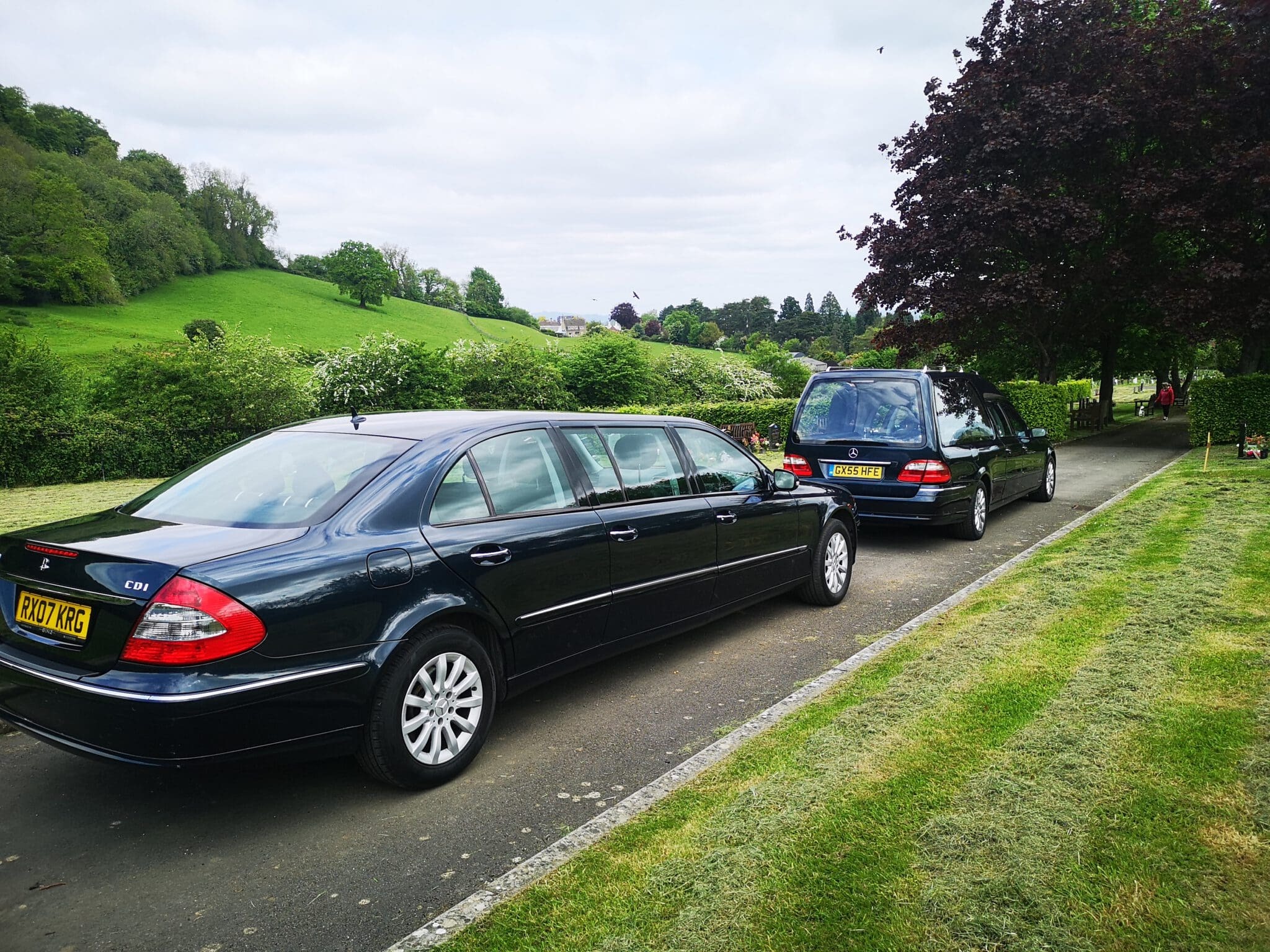 Two black funeral cars are parked on a narrow road beside a lush, green lawn and trees, with rolling hills and cloudy sky in the background. Both have yellow license plates, and one car has tinted windows.