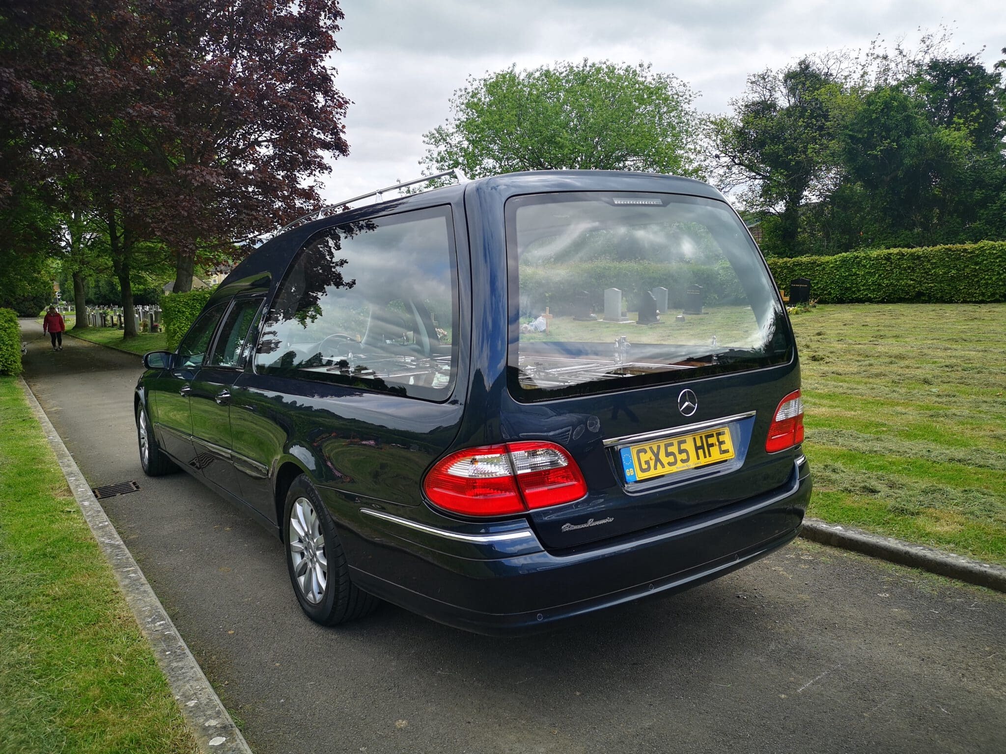 A black Mercedes-Benz hearse is parked on a paved path next to a grassy cemetery, with gravestones visible in the background under cloudy skies.