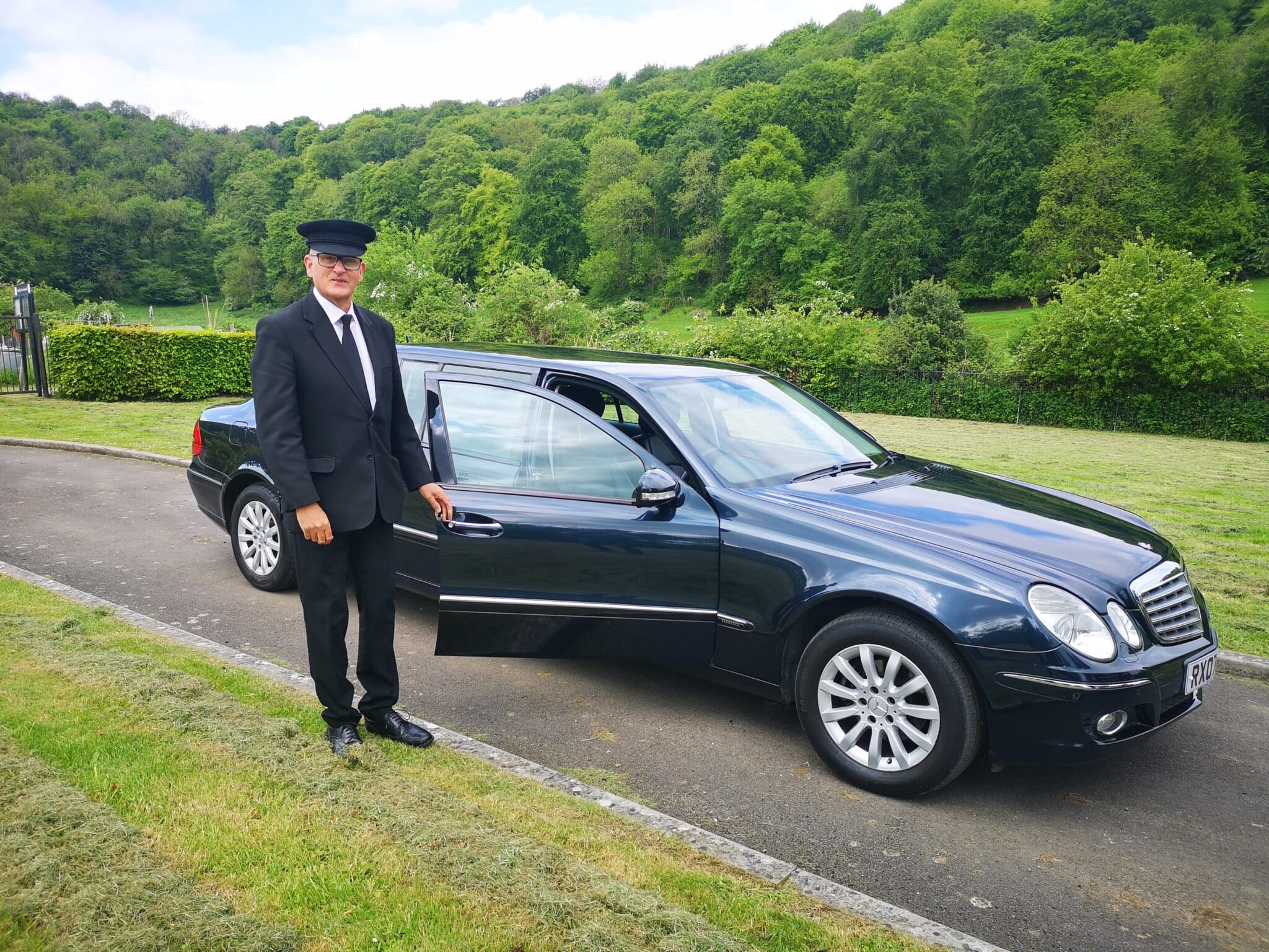A chauffeur in a black suit, tie, and cap stands by an open door of a black sedan parked on a paved road, with green trees and grass in the background.
