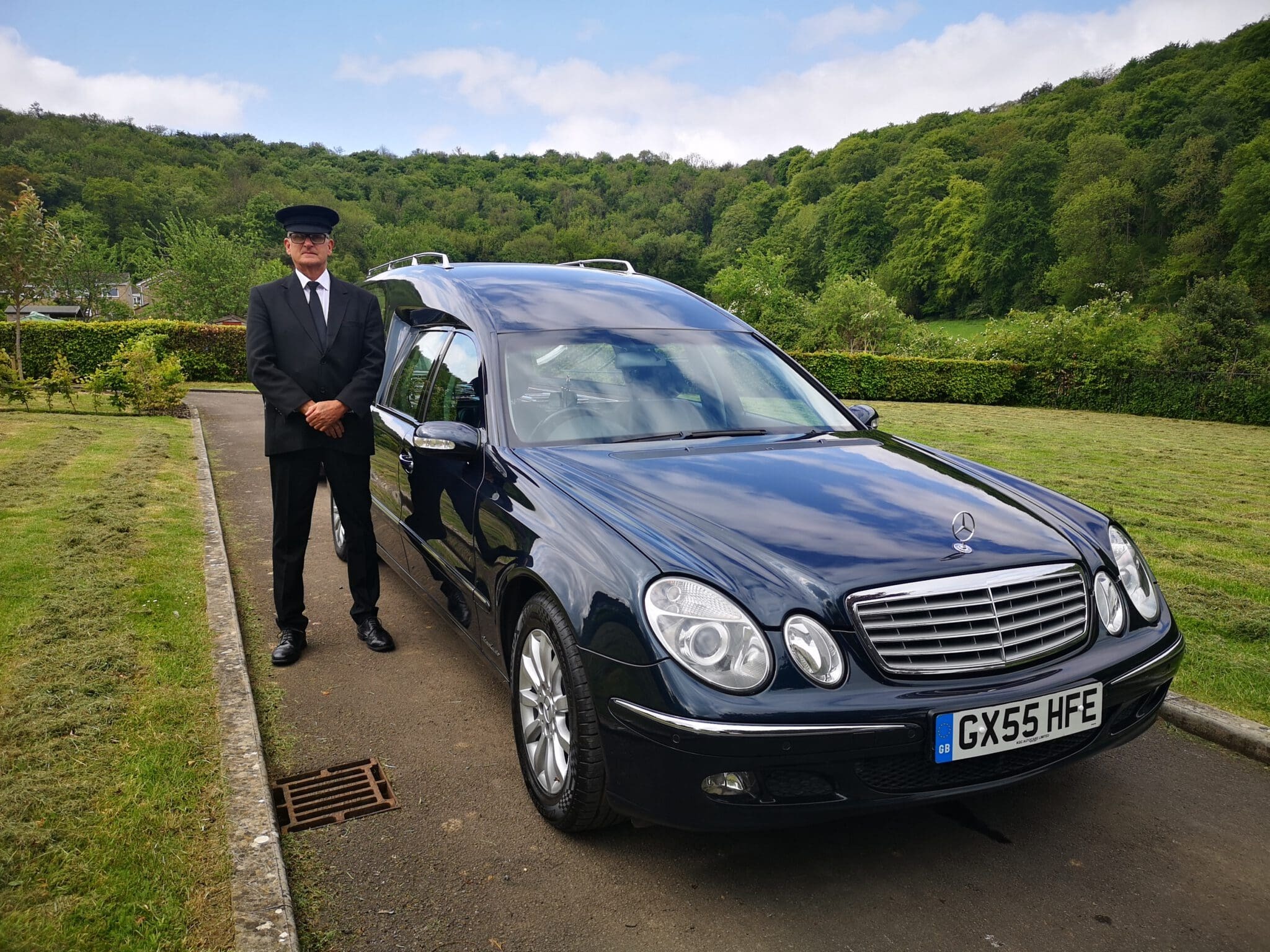 A chauffeur in a black suit and cap stands next to a black Mercedes-Benz hearse with a UK license plate, parked on a paved driveway with green lawns and trees in the background.
