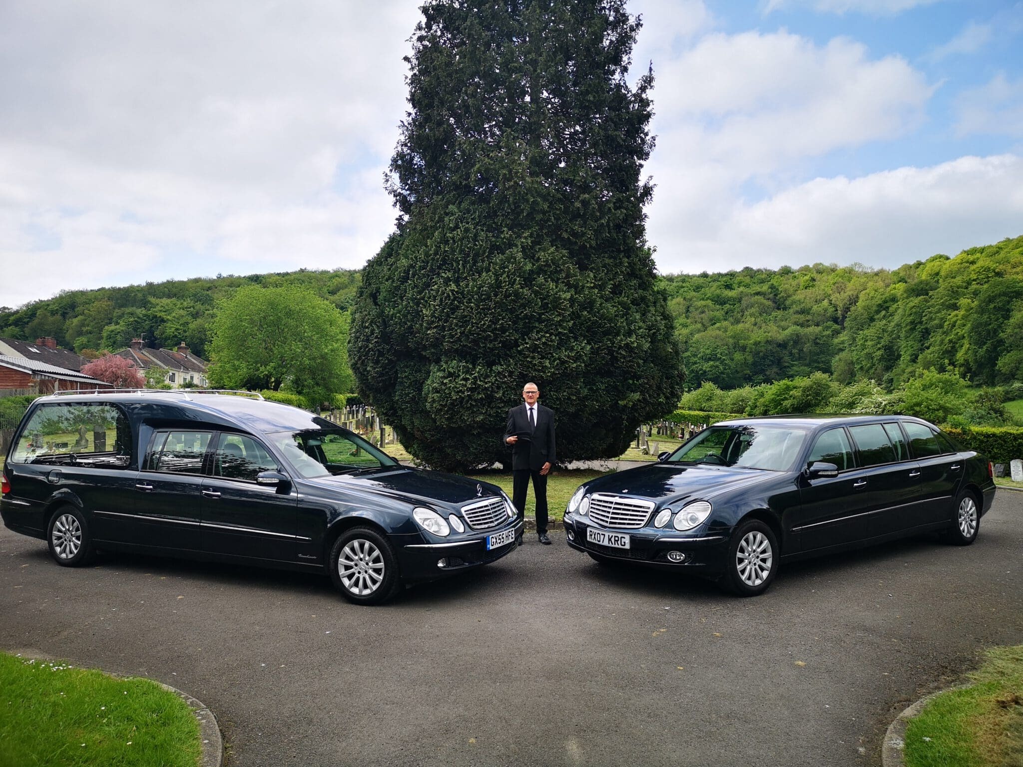 A man in a suit stands between a black hearse and a black limousine parked on a driveway, with a large tree and green landscape in the background.