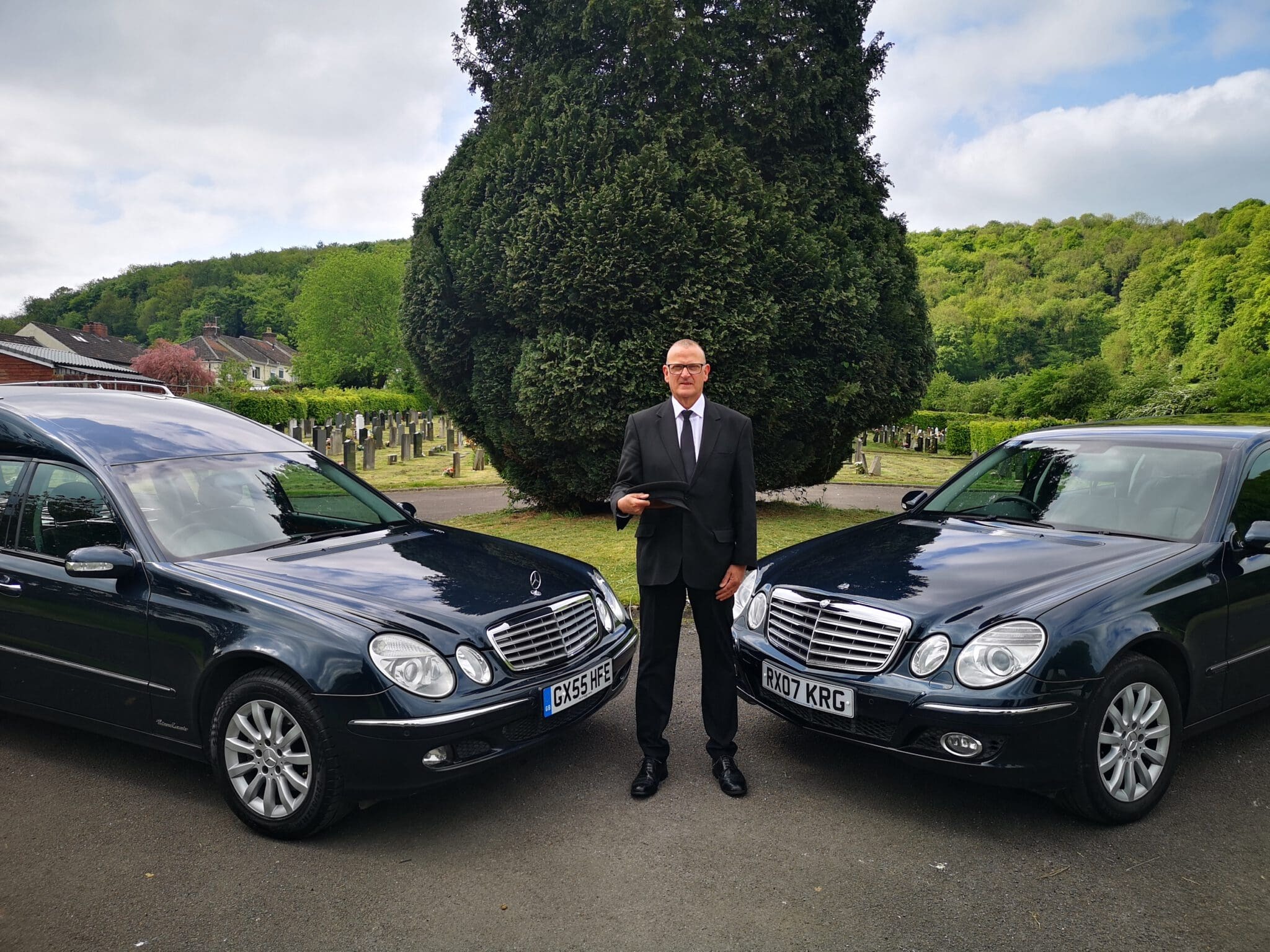 A man in a black suit stands between two black funeral cars in front of a cemetery with green trees and hills in the background, holding a folder and looking at the camera.