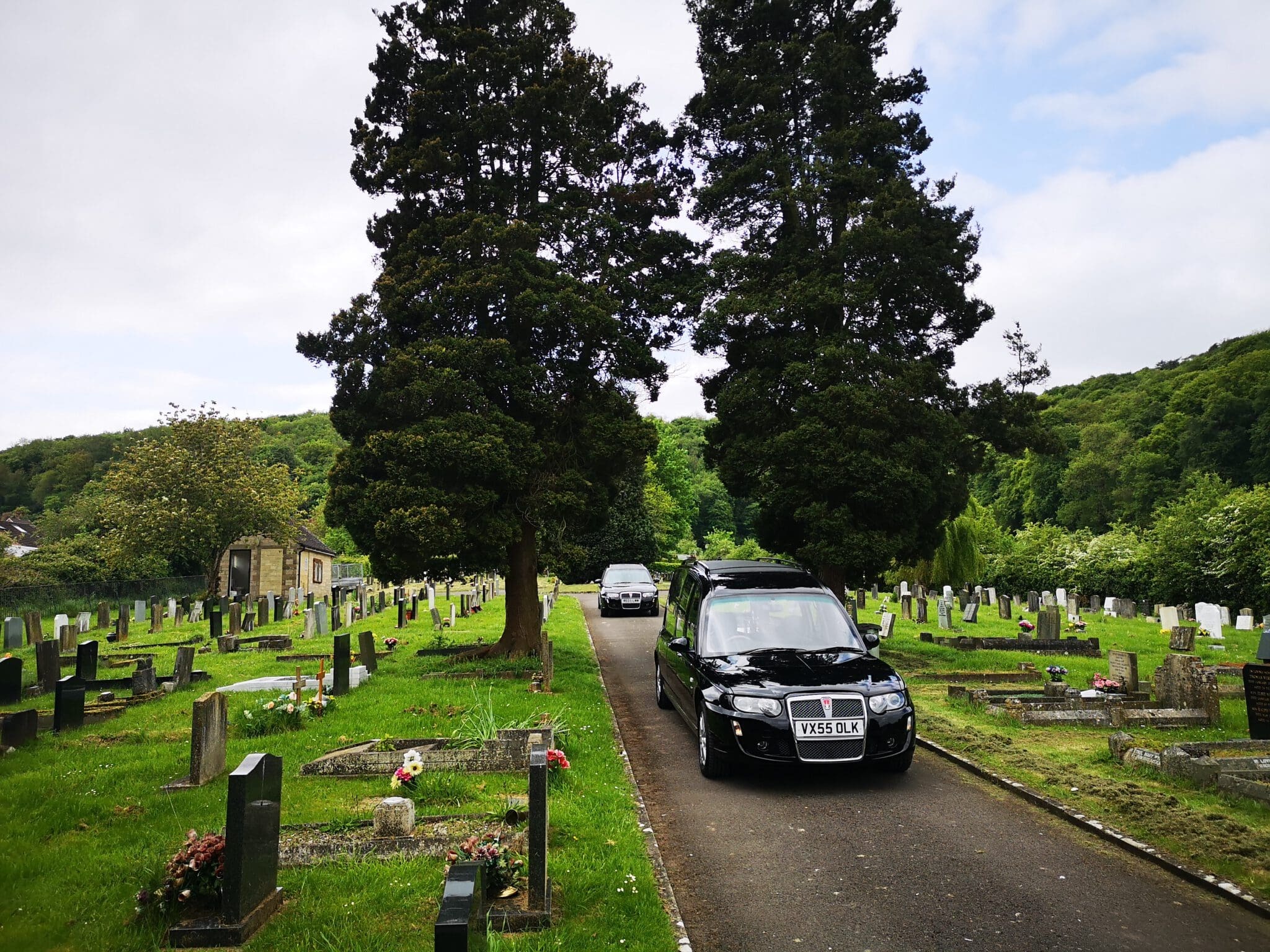 A black hearse and another car drive down a path through a cemetery with headstones and tall trees, surrounded by green grass and rolling hills under a partly cloudy sky.