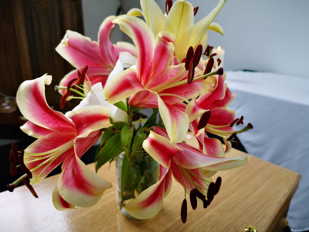 A clear glass vase holds a bouquet of vibrant pink and yellow lilies with dark stamens, placed on a wooden surface in a softly lit room.