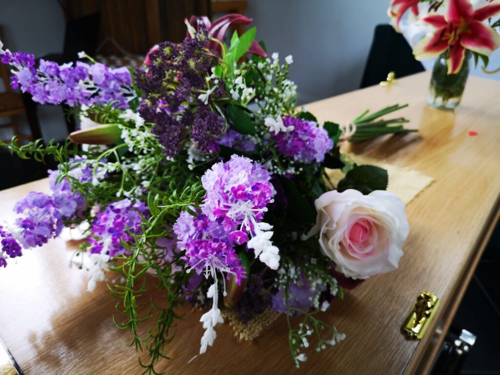 A bouquet of purple, pink, and white flowers rests on a light wooden coffin, with another floral arrangement blurred in the background.
