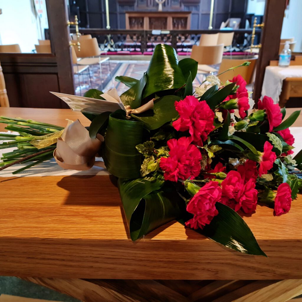 A bouquet of red flowers and green leaves rests on a wooden surface inside a church, with chairs, an altar, and other furniture visible in the background.